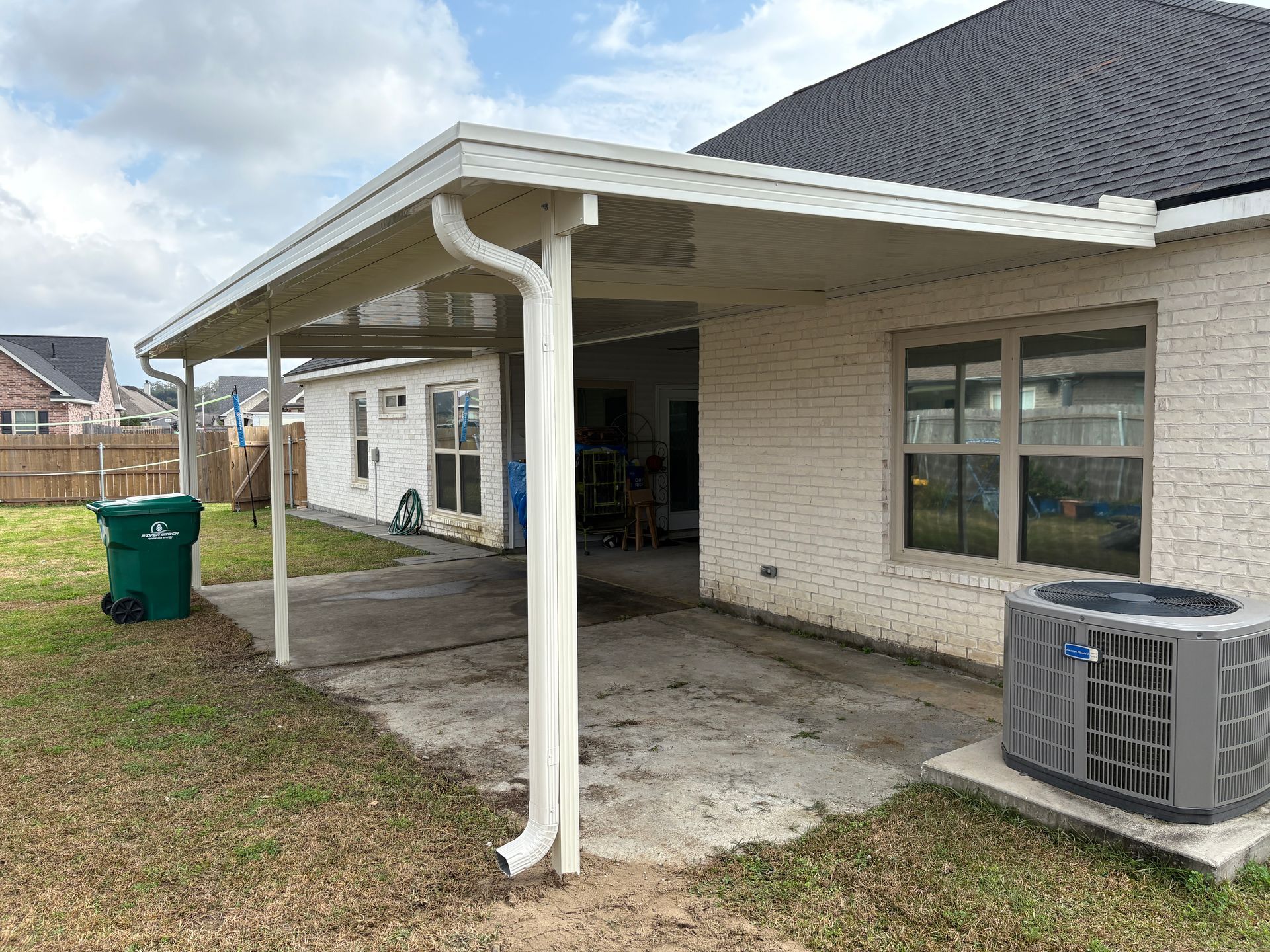 A white brick house with a covered concrete patio, a central support column, a gutter system, and an outdoor AC unit.