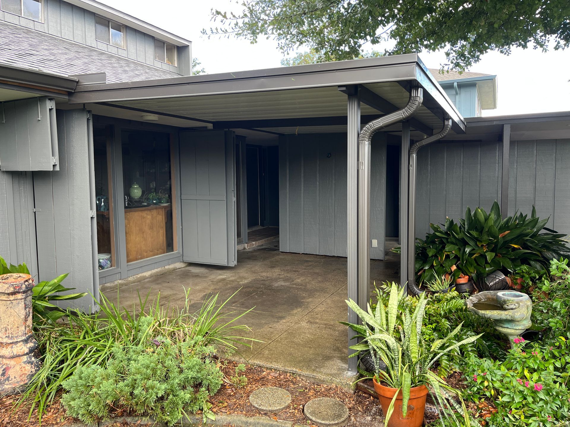 A covered gray patio with a concrete floor, adjacent to a gray house with wood-paneled walls and lush green plants.