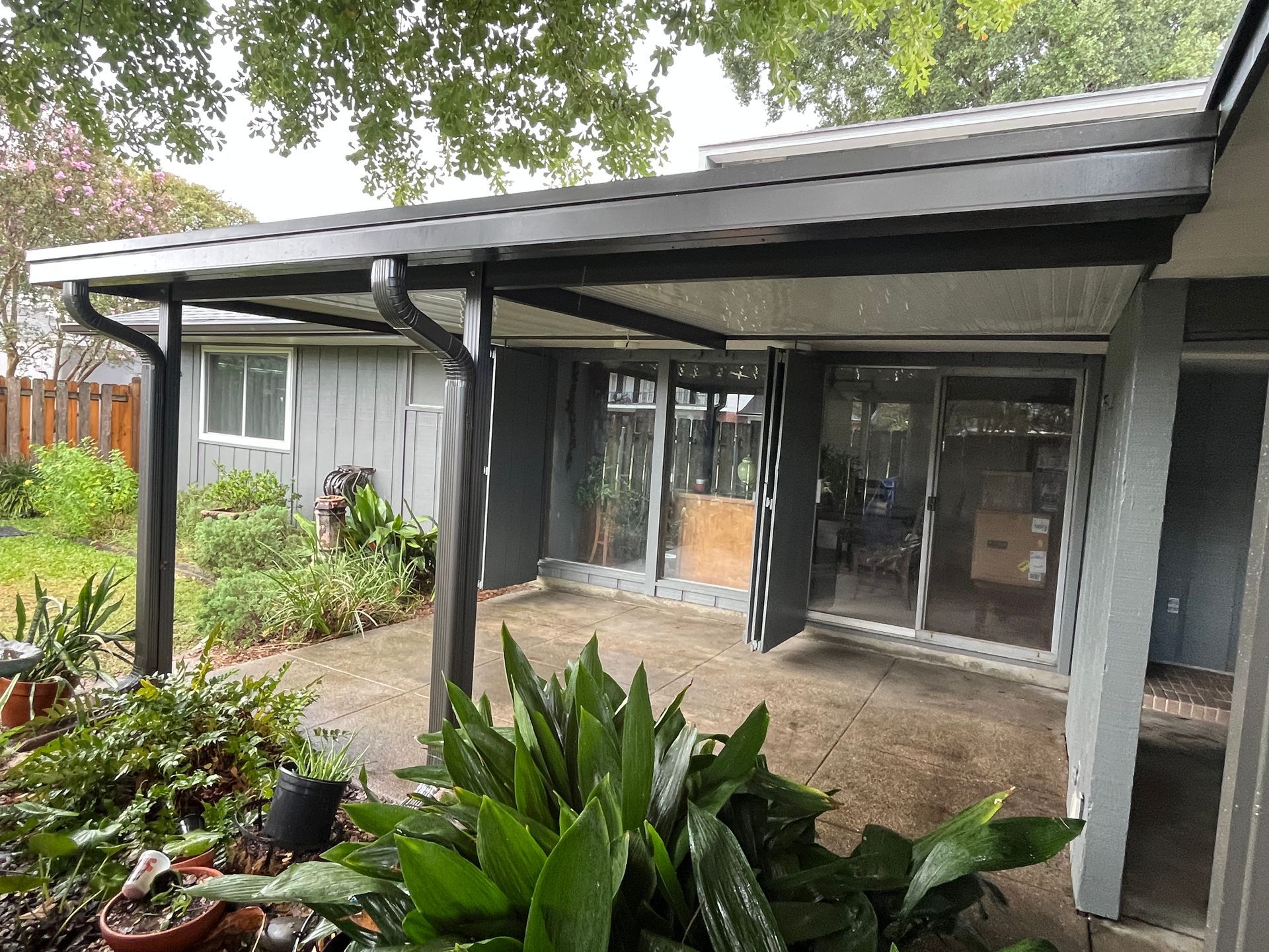 A dark gray, covered patio attached to the back of a house, featuring sliding glass doors and lush green plants.