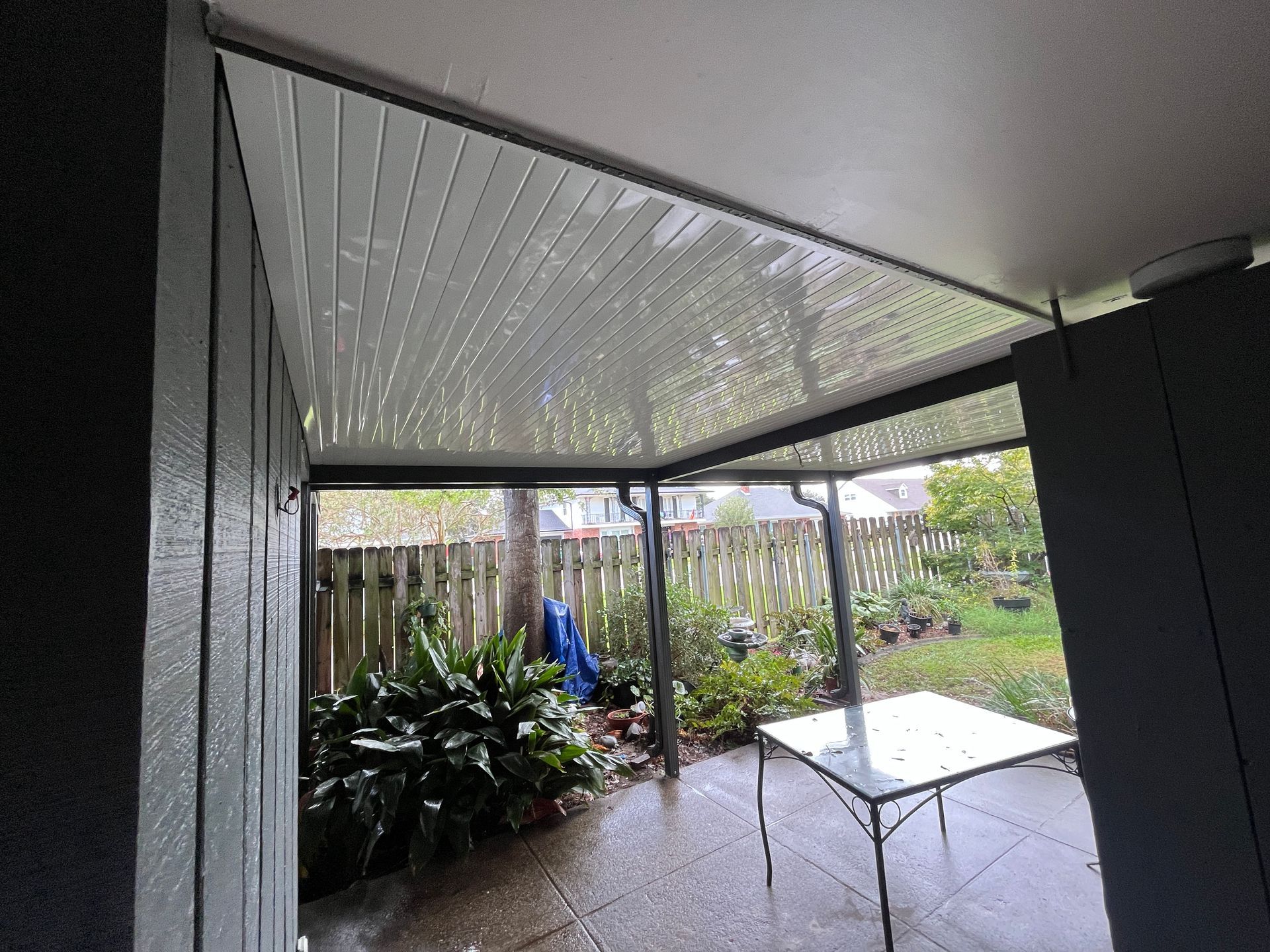A covered patio with a white corrugated ceiling, metal support posts, a small glass table, and a backyard garden.