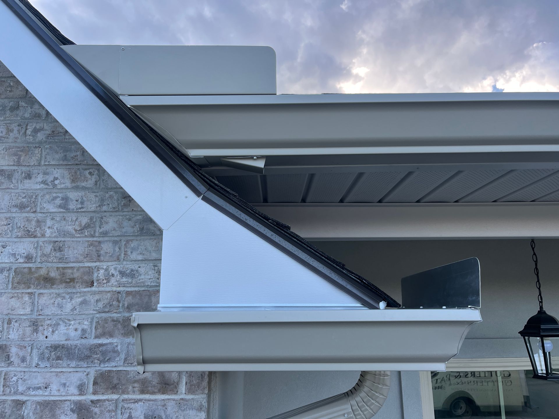 A close-up view of a roof eave showing light-colored gutters, a white fascia board, and tan brick siding.