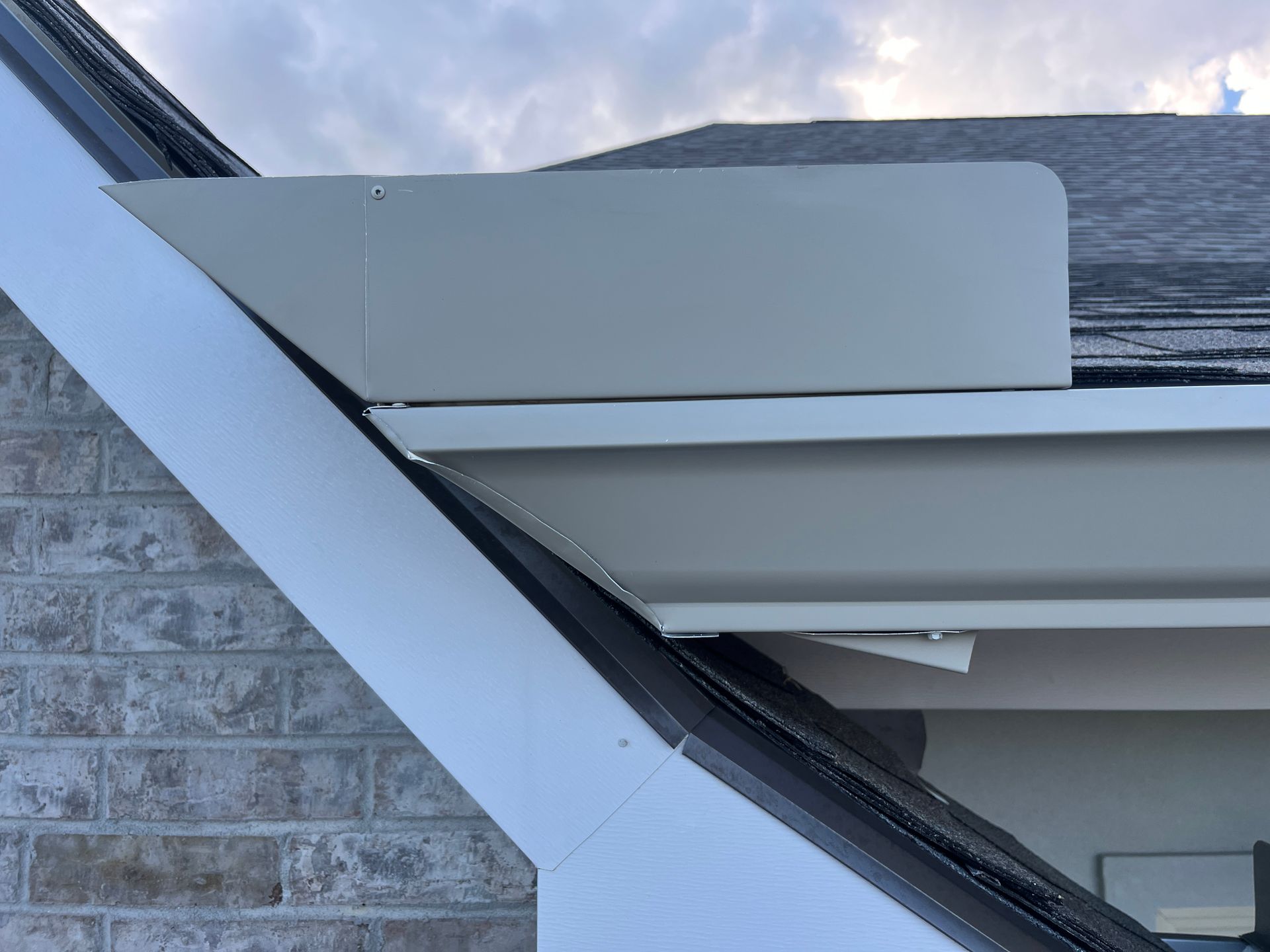 A side view of a light-colored metal roof edge and gutter system installed against a brick wall and sloped roof.