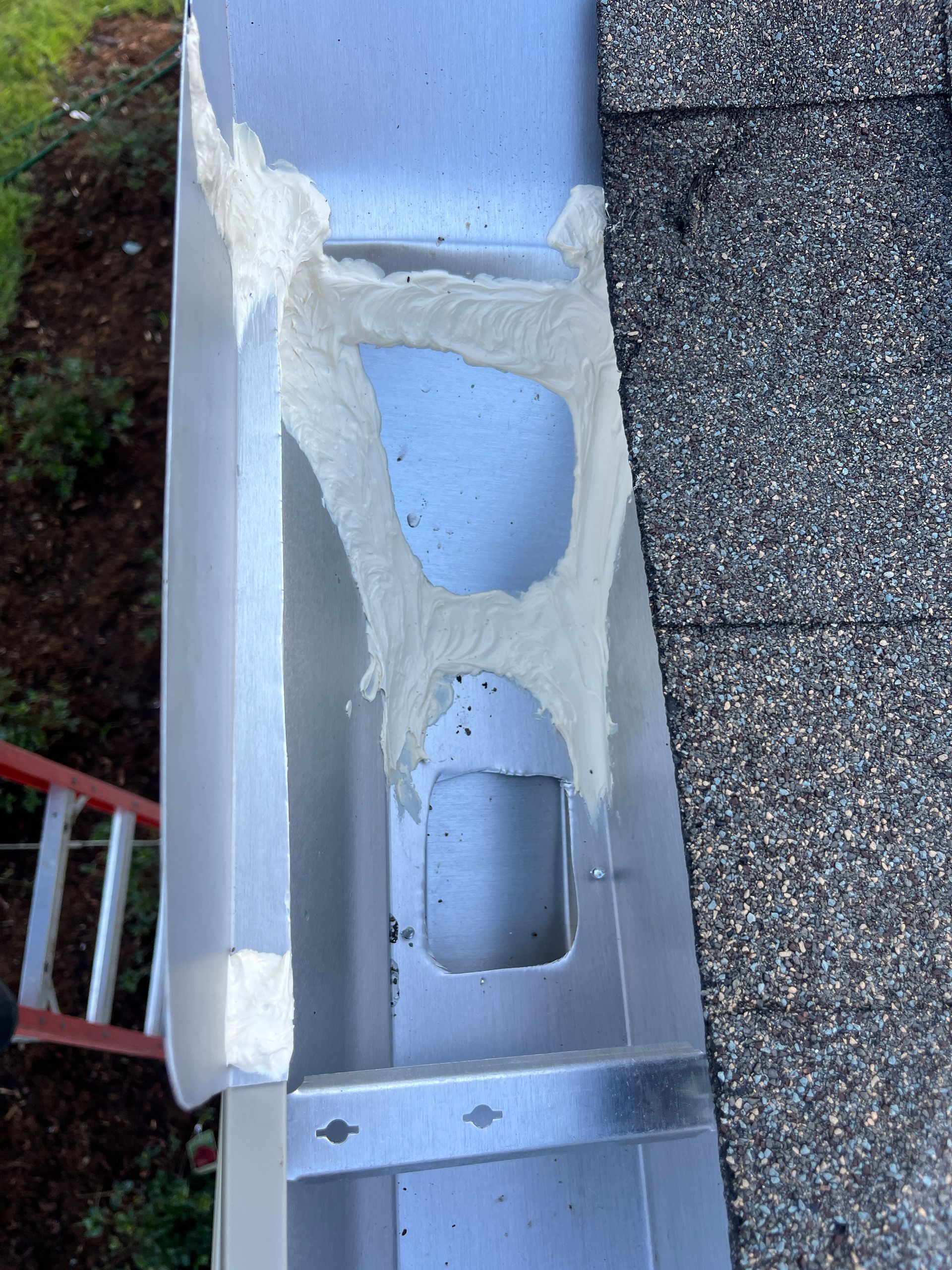 A close-up view of a metal rain gutter containing large gaps filled with messy, light-colored spray foam sealant.