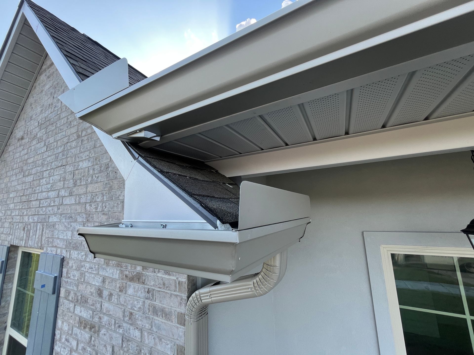 A beige house exterior showing a corner gutter section with a protective cover and a metal downspout.