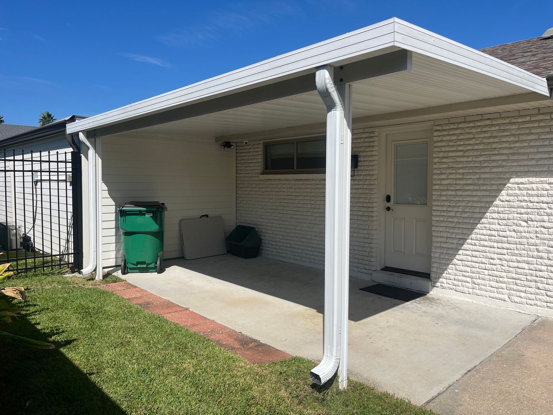 A white brick house exterior with a covered patio, a green trash bin, and a white downspout on a sunny day.
