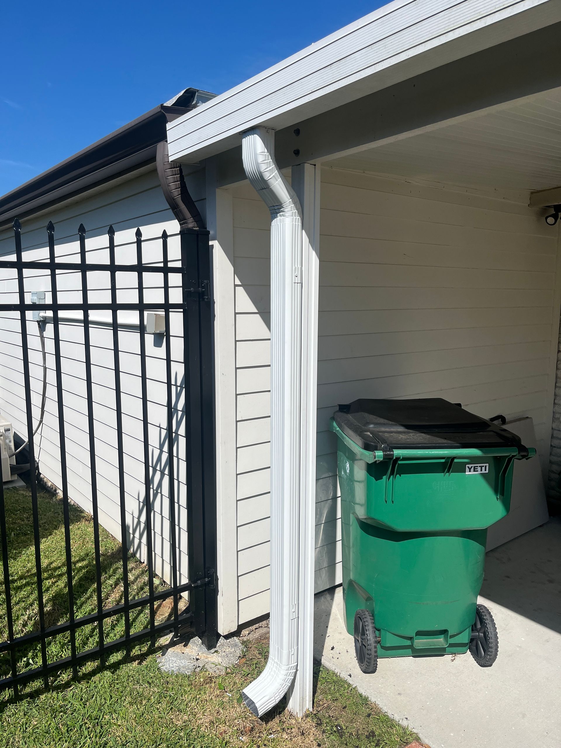 A white downspout mounted on a beige wall next to a black metal fence and a green trash bin outside a home.