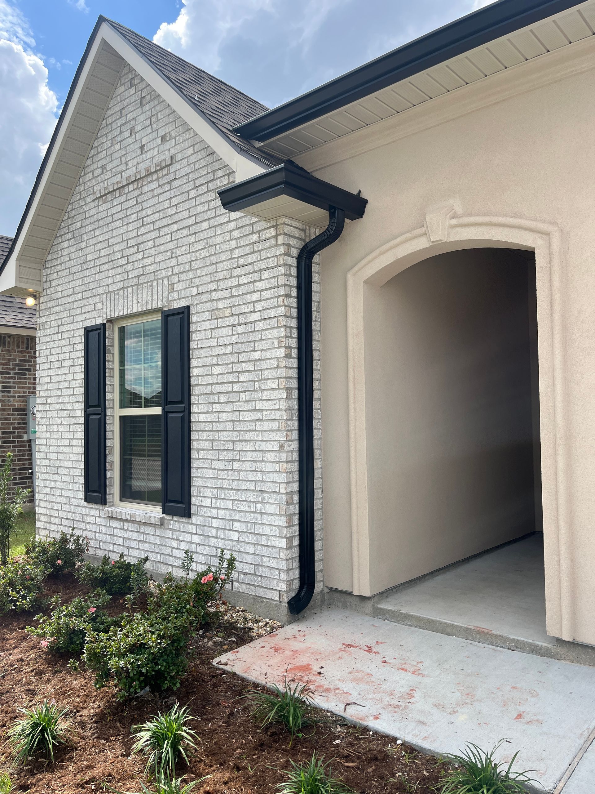 A house exterior with light gray brick, cream stucco, black shutters, and a black downspout near the recessed entryway.