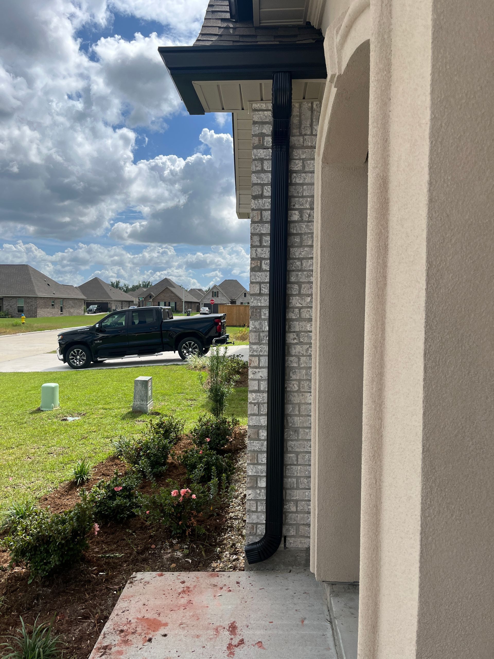 A black downspout attached to the corner of a brick and stucco house exterior, near a landscaped front yard.