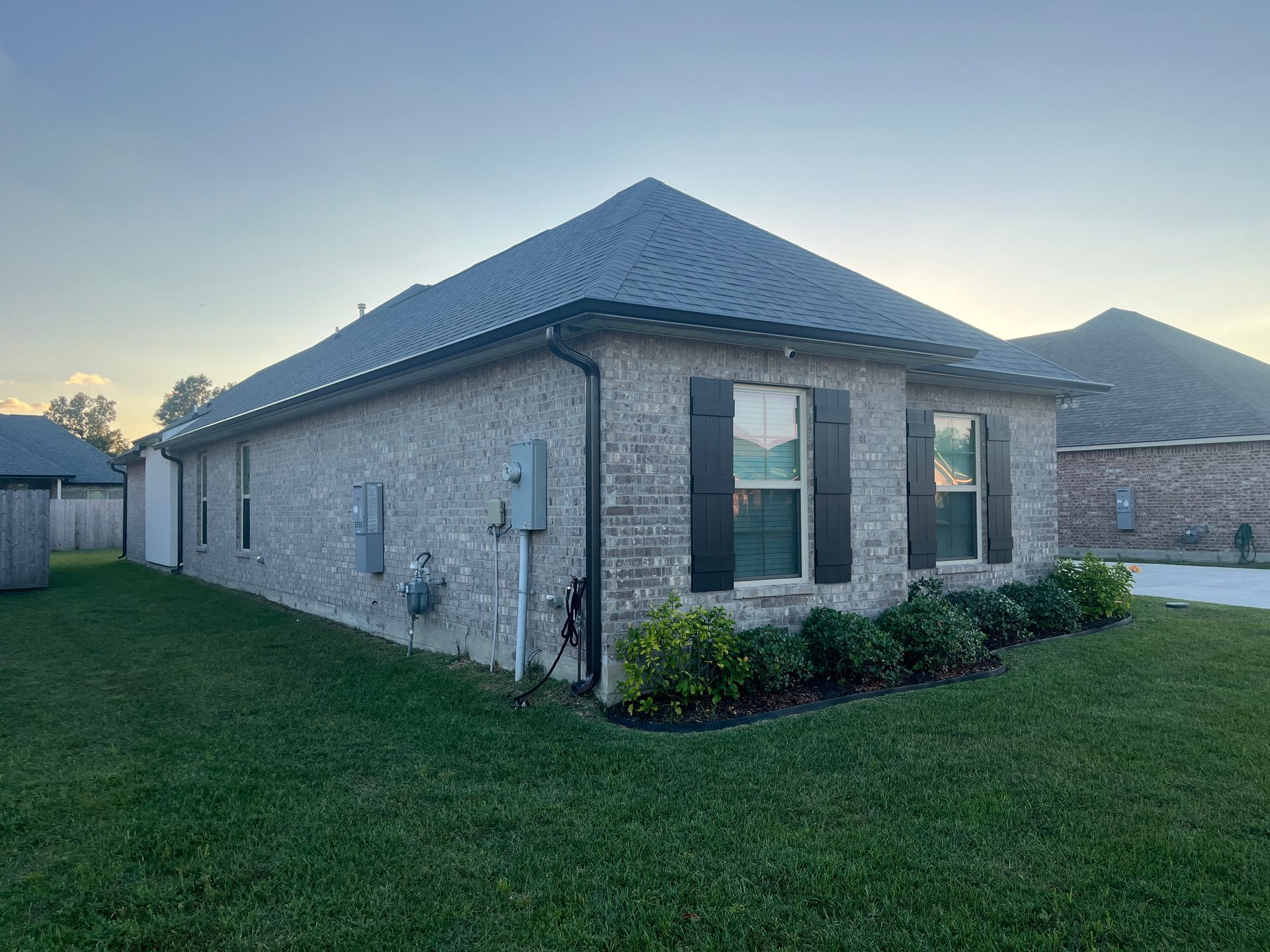 A single-story, light-colored brick house with a dark shingled roof, black window shutters, and a lawn under a blue sky.