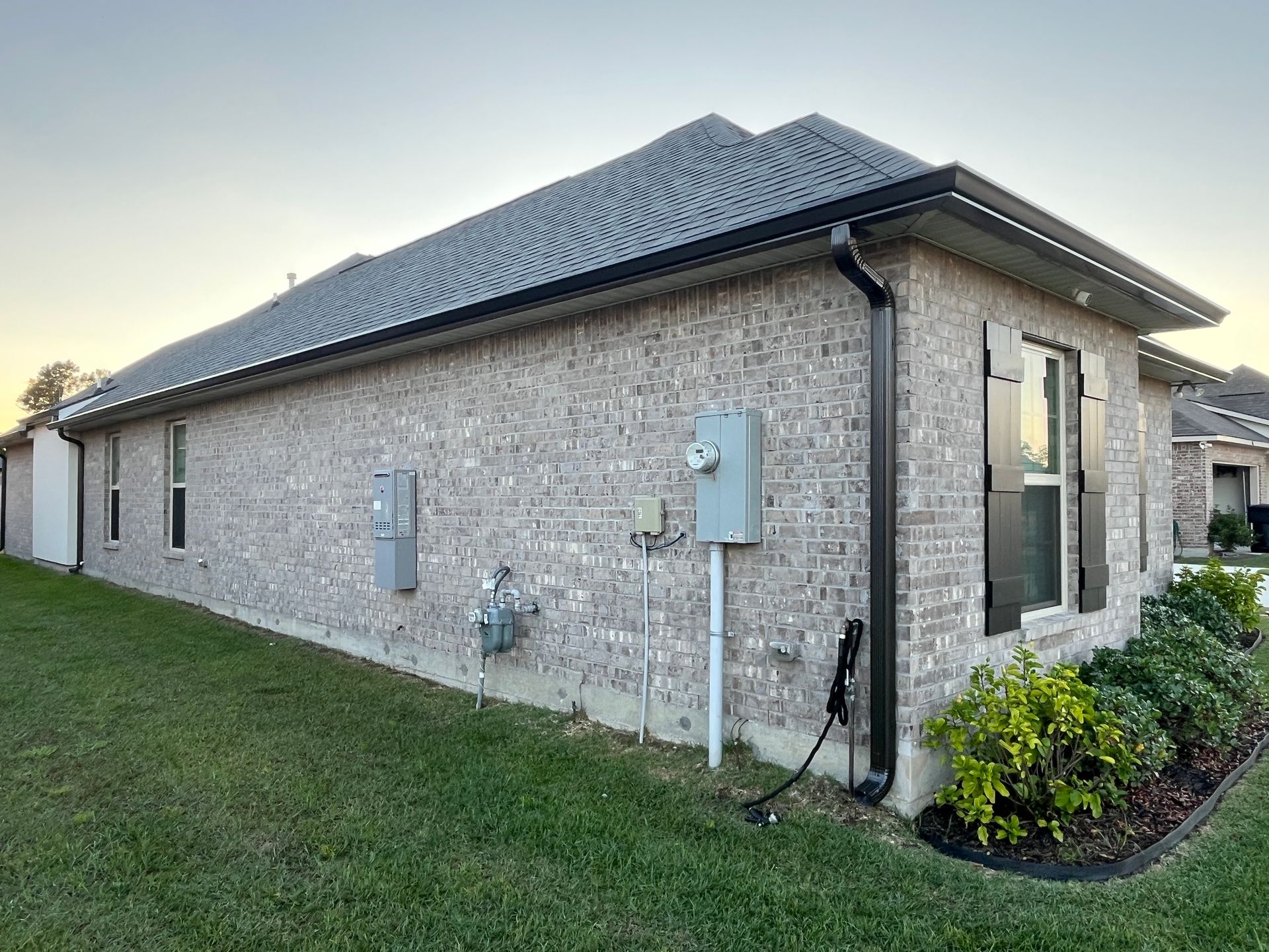 A side view of a modern house with grey brick siding, a dark shingle roof, black shutters, and utility boxes.