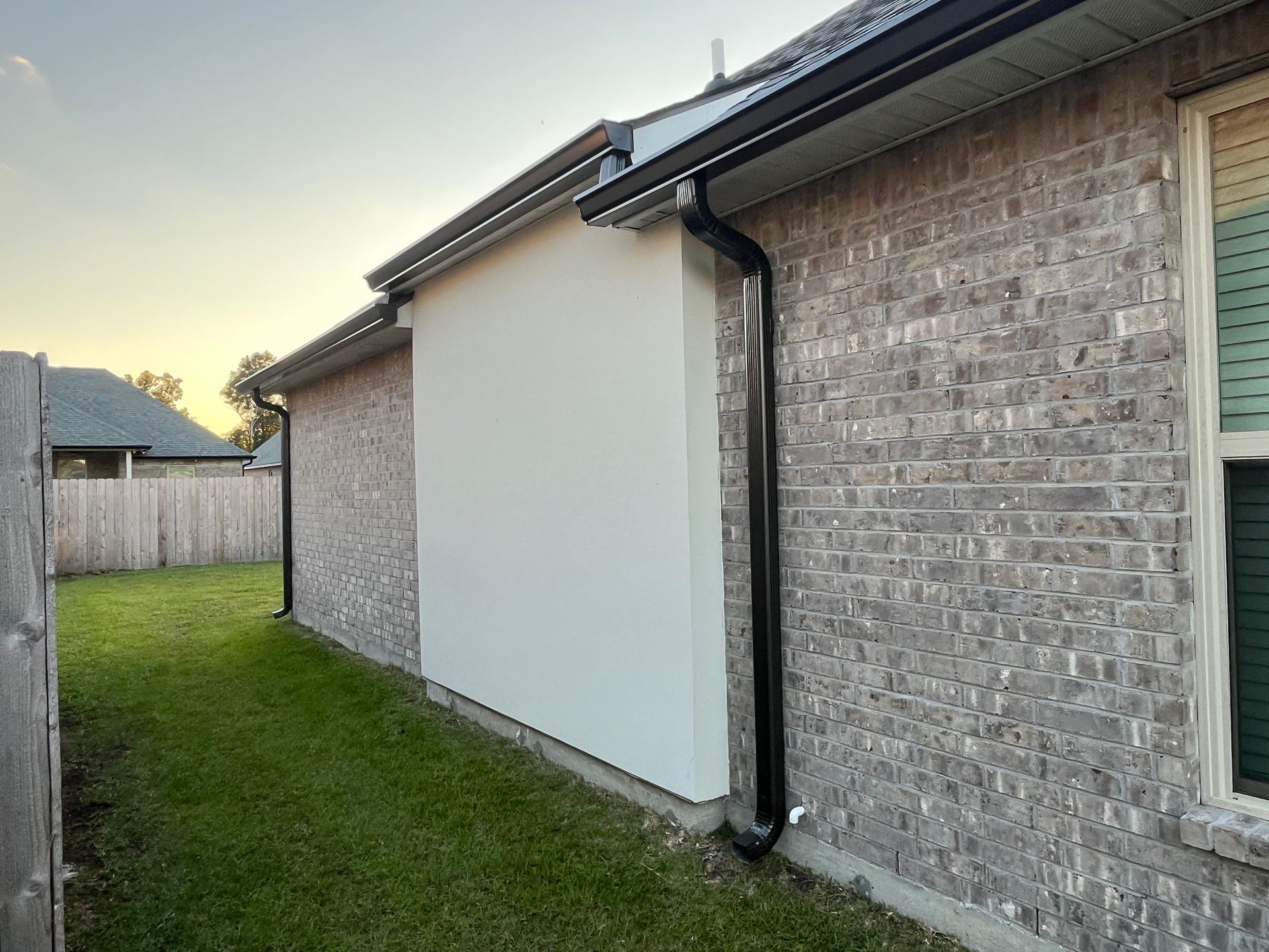 A side view of a brick house with a large, smooth, light-colored stucco panel and a black vertical gutter downspout.