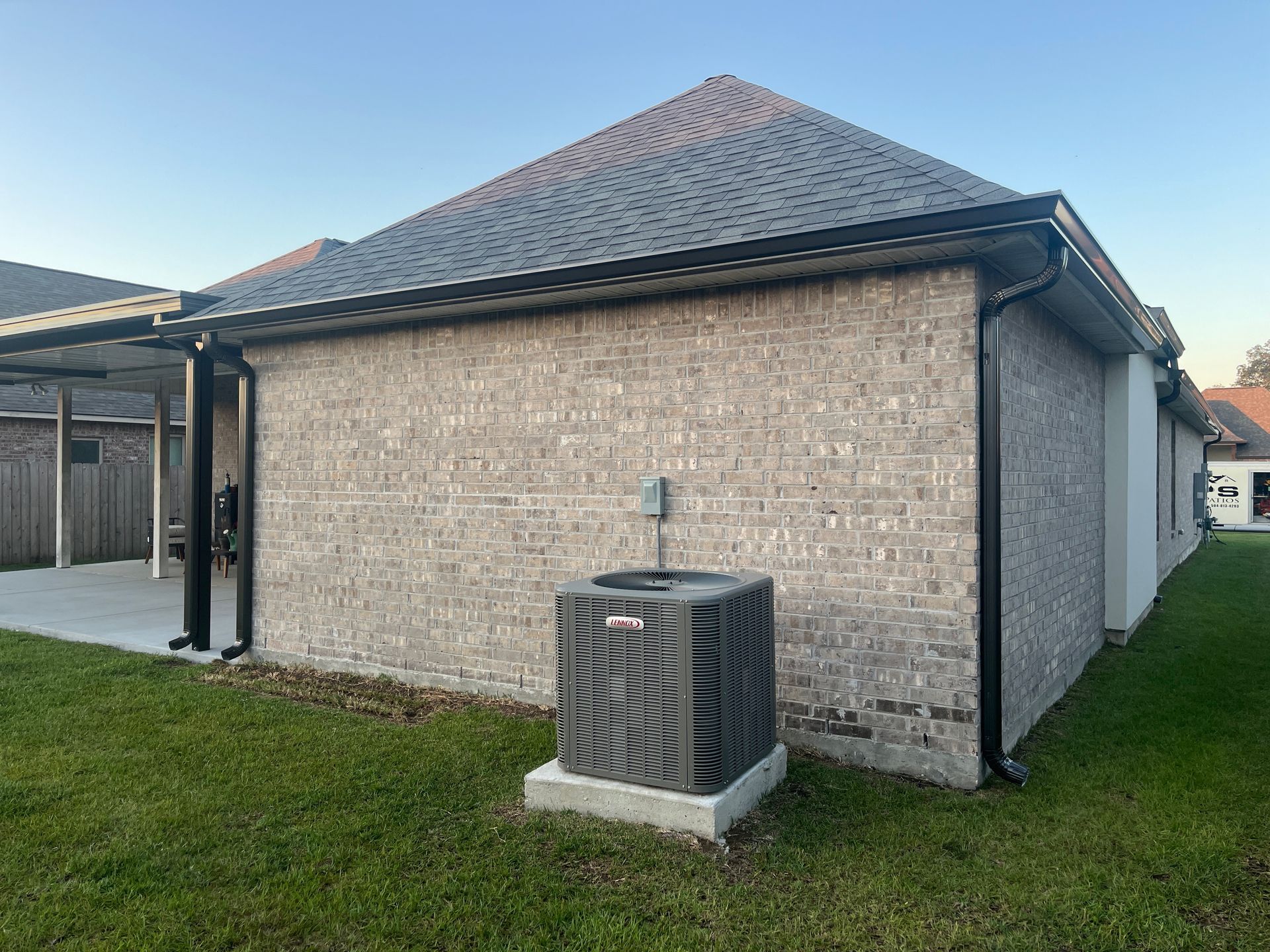 A brick exterior wall of a house with a grey HVAC unit on a concrete pad, a black gutter, and a side patio.