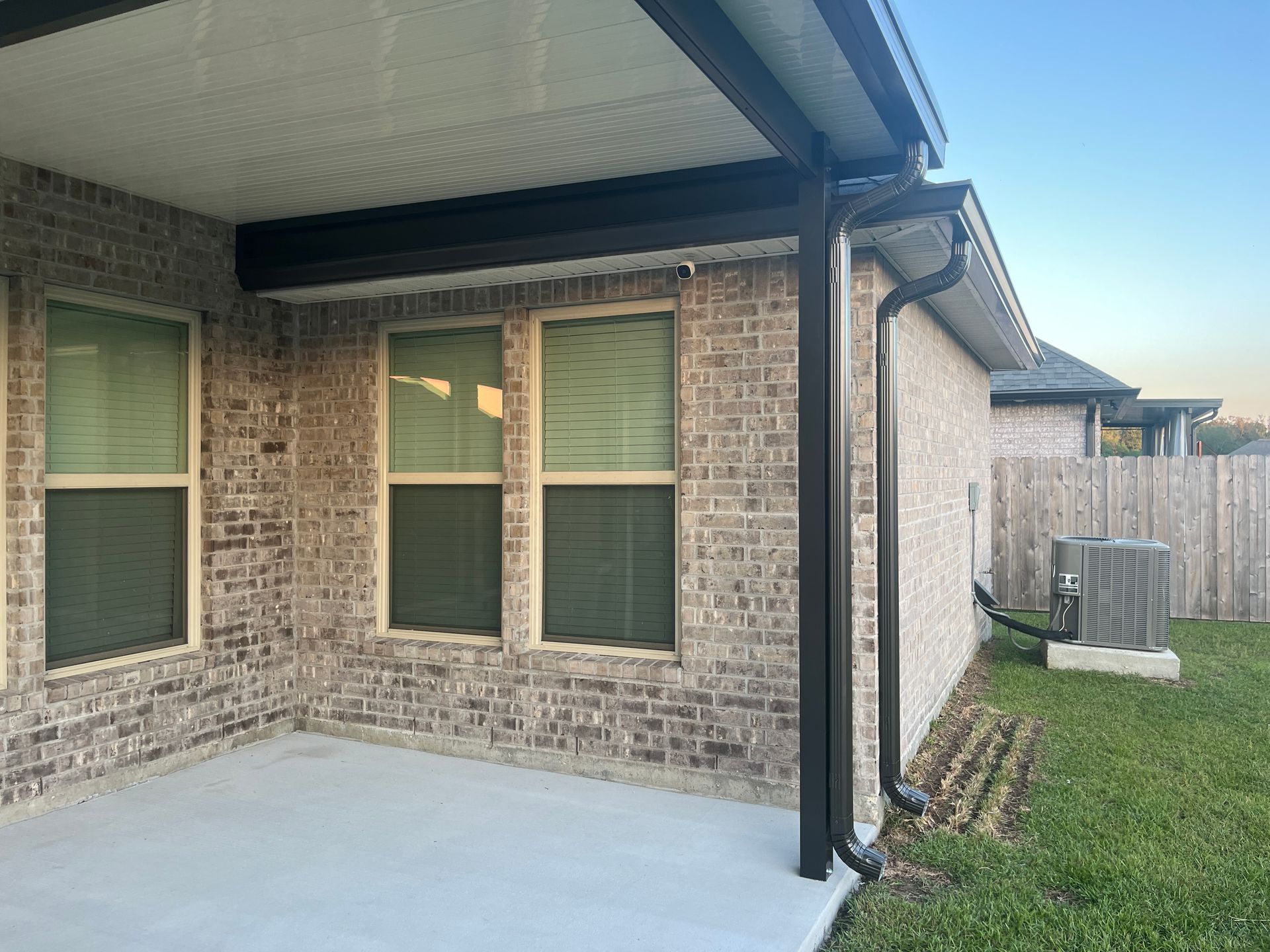 A beige brick exterior of a house featuring a covered patio, two windows with blinds, and a black downspout.