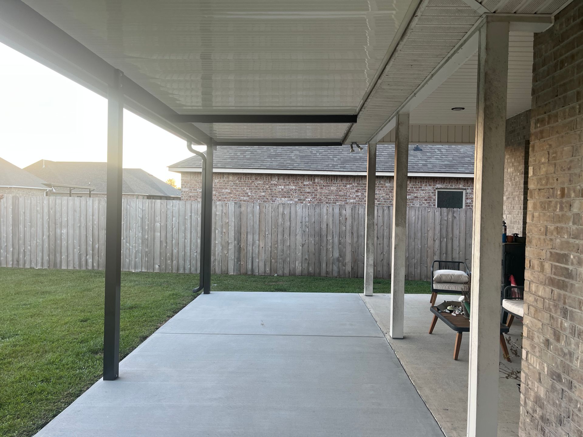 A covered patio with a concrete floor, wooden support beams, and a view of a grassy backyard and a wooden fence.