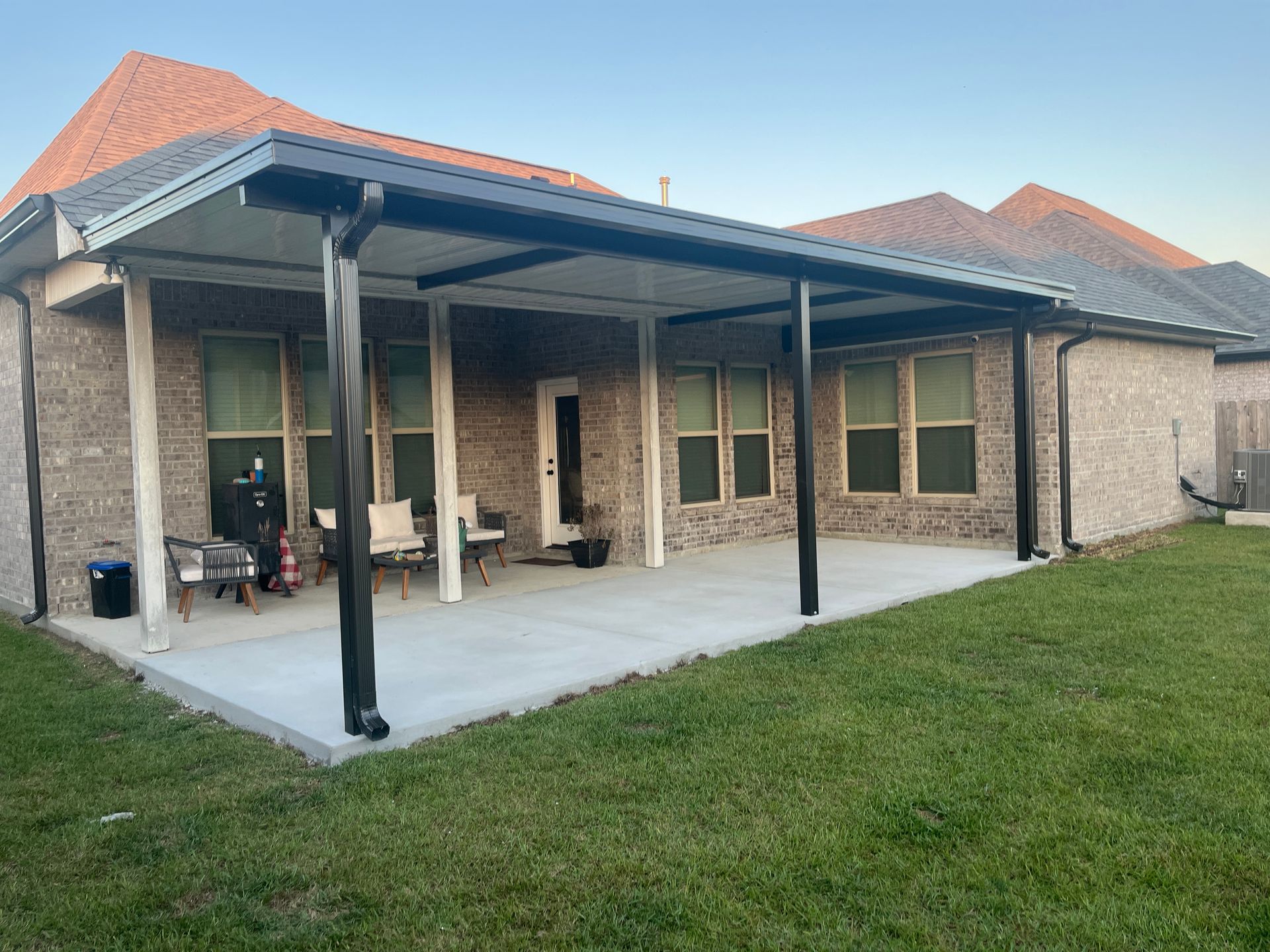 A back patio with a dark metal-framed covered roof, concrete flooring, and outdoor furniture against a brick house.