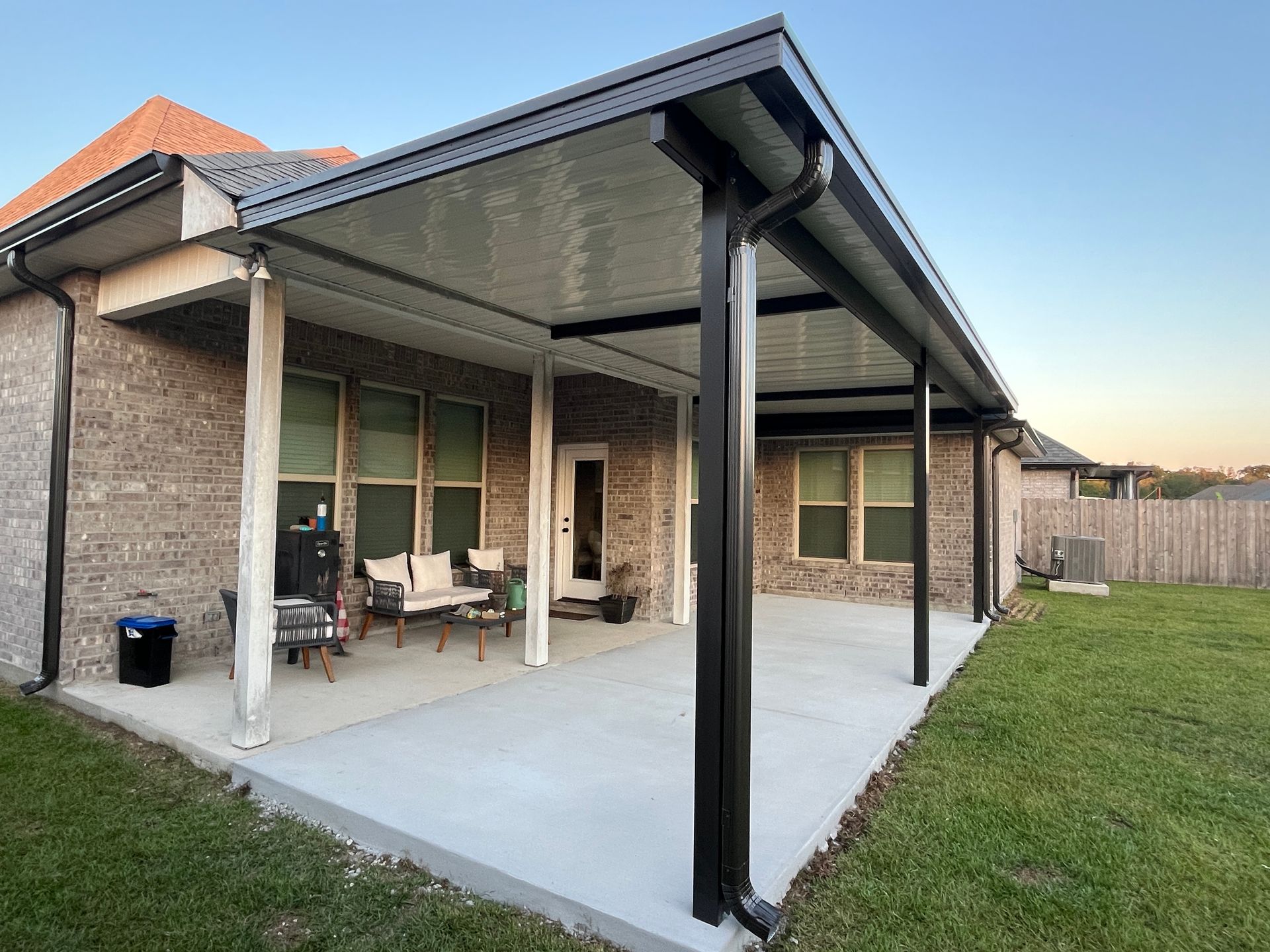 A covered outdoor patio with a concrete slab, black support columns, and white seating against a brick house wall.