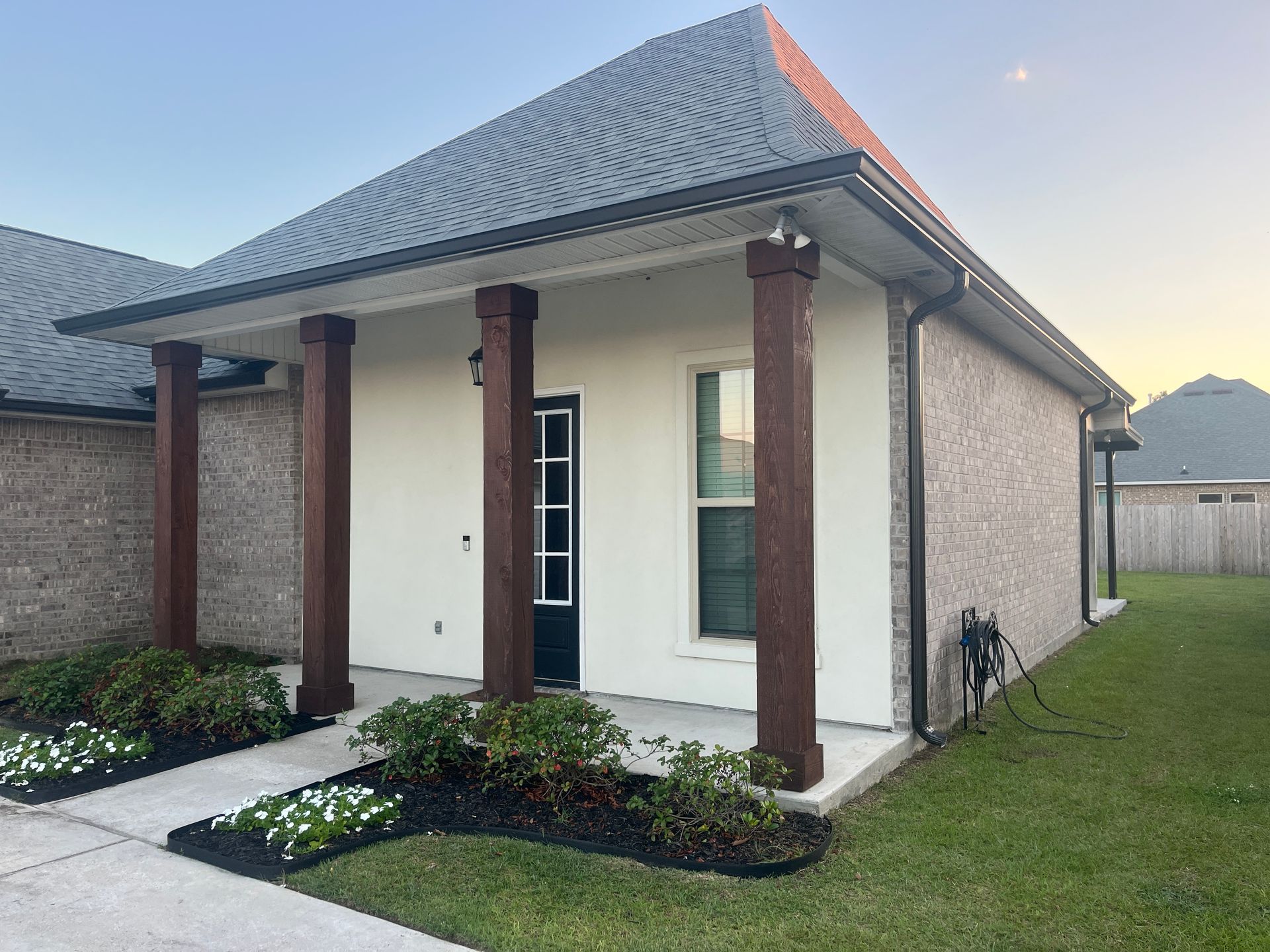 A single-story brick house with a covered front porch featuring three dark wooden pillars, a black door, and landscaping.