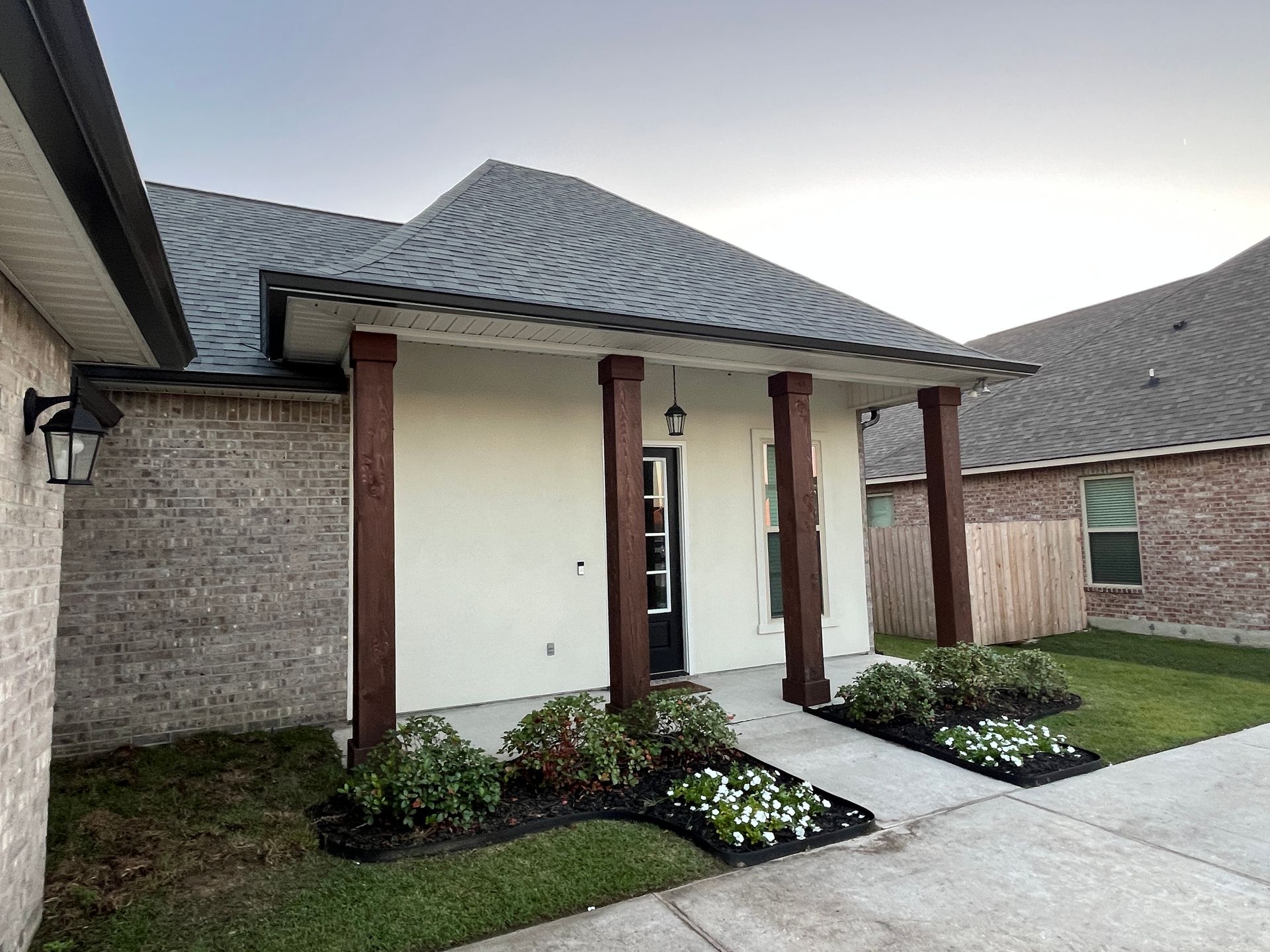 A house exterior with a covered entryway, four dark wooden posts, white stucco, brick walls, and small landscaped bushes.