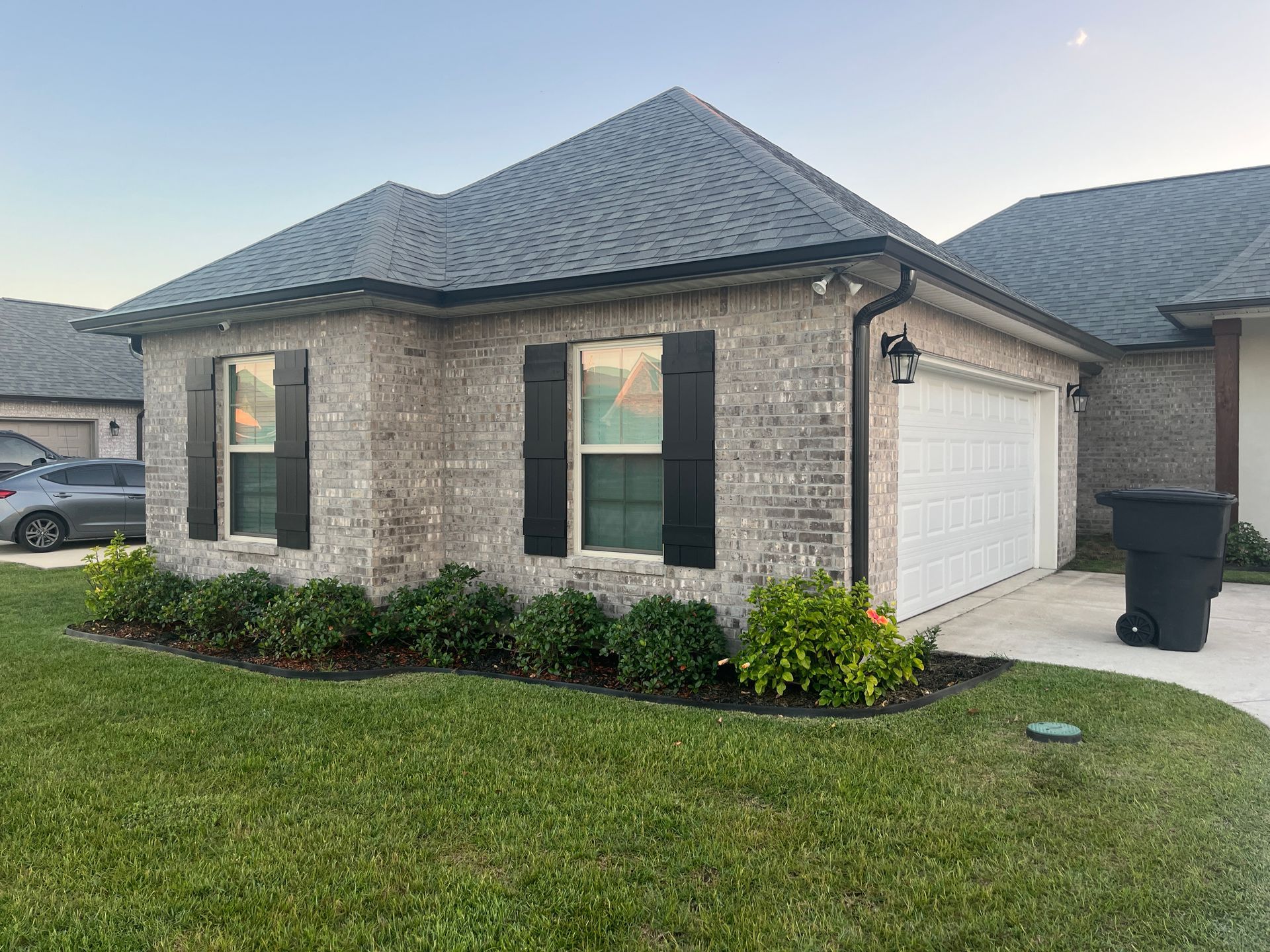 A tan brick single-story house with a grey shingled roof, black shutters, and a one-car garage, seen from the front lawn.