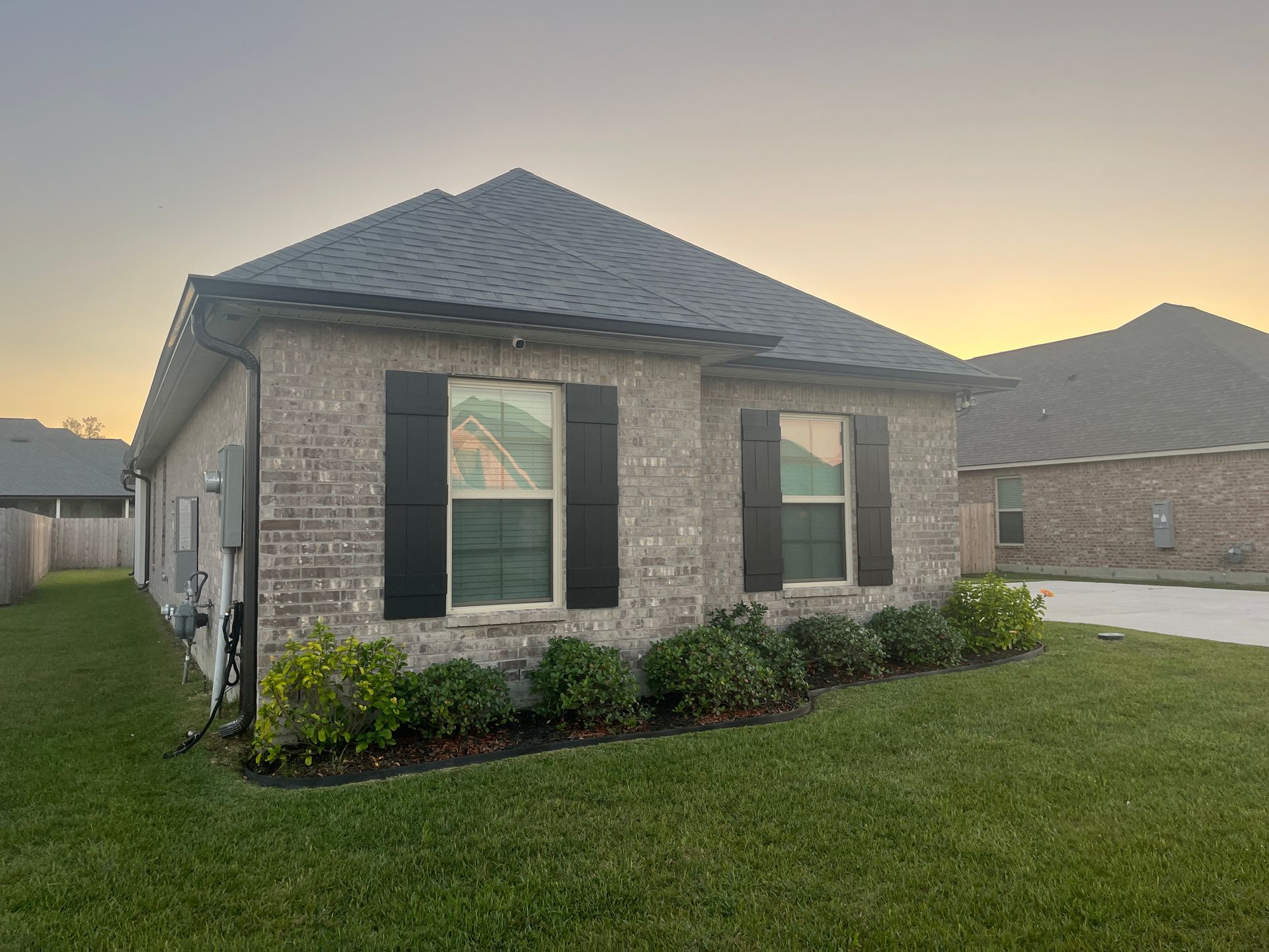 A tan brick, single-story house with a dark roof and black window shutters, surrounded by green grass at sunset.