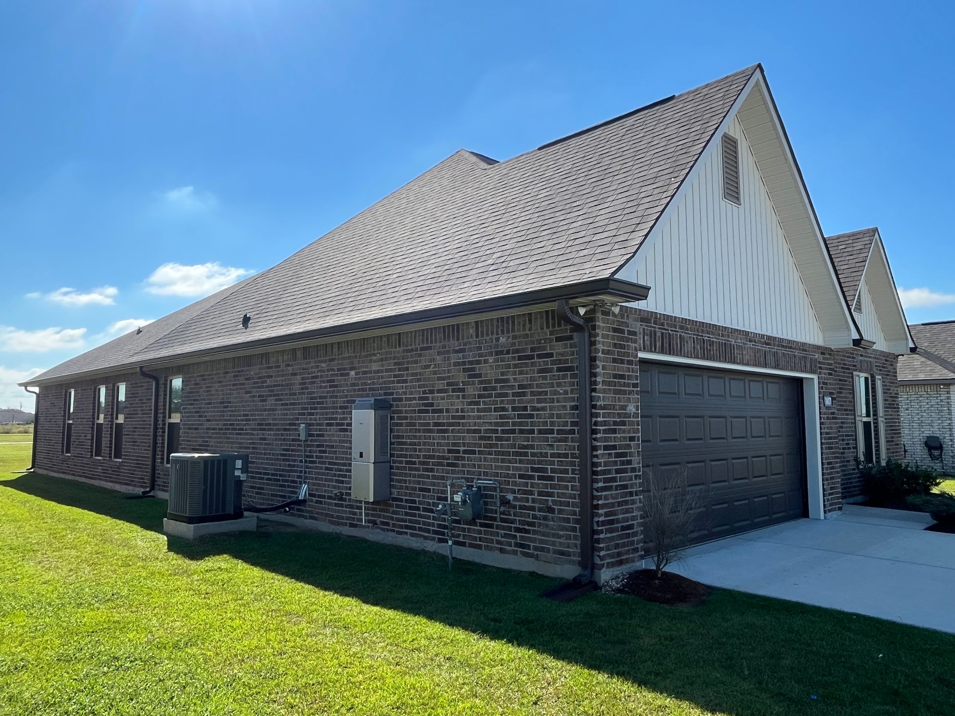 A side view of a modern brick house with a brown shingled roof, an attached garage, and a lush green lawn under a blue sky.