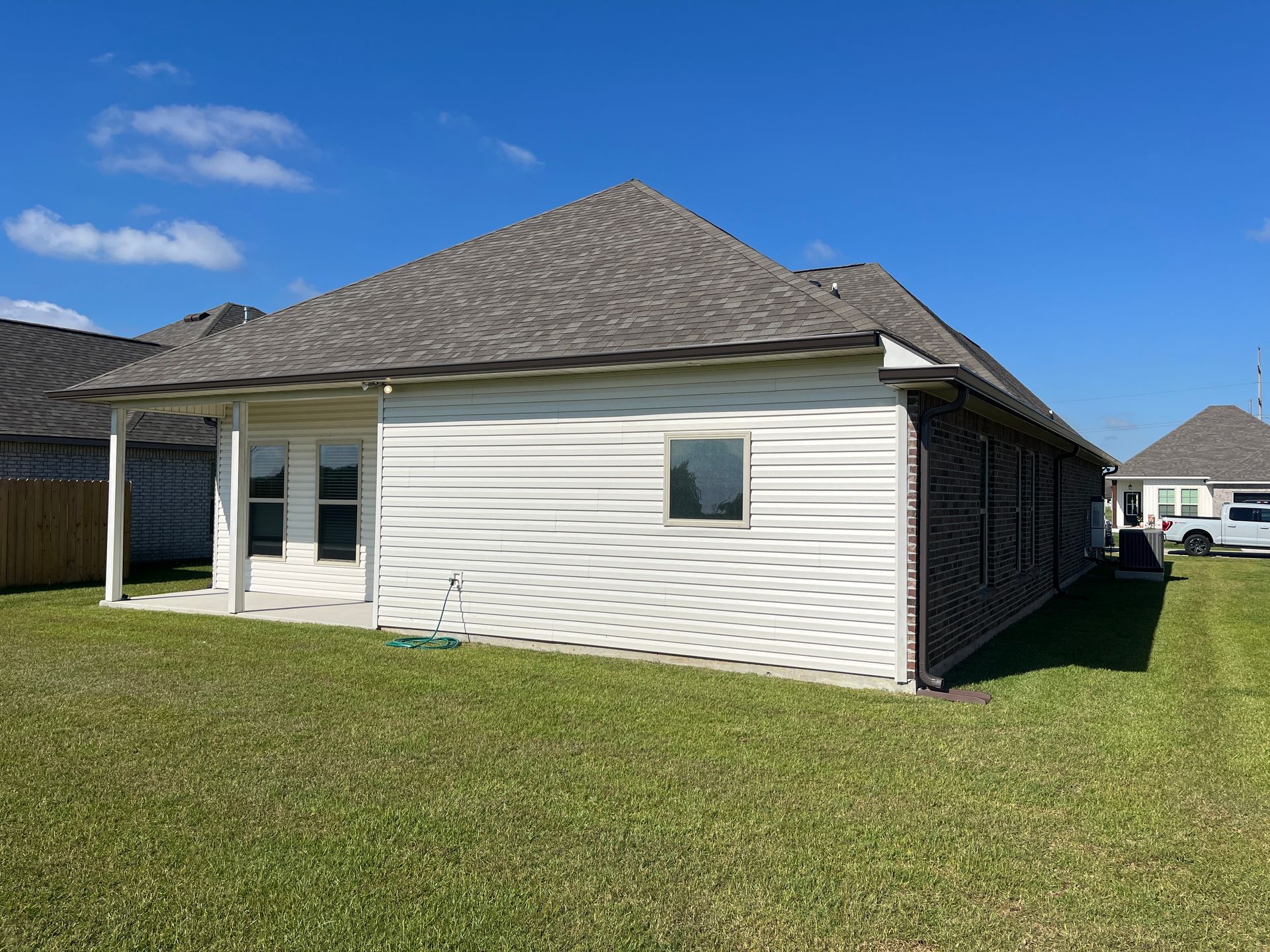 Backyard view of a single-story house with cream siding, dark brick trim, and a covered patio under a blue sky.