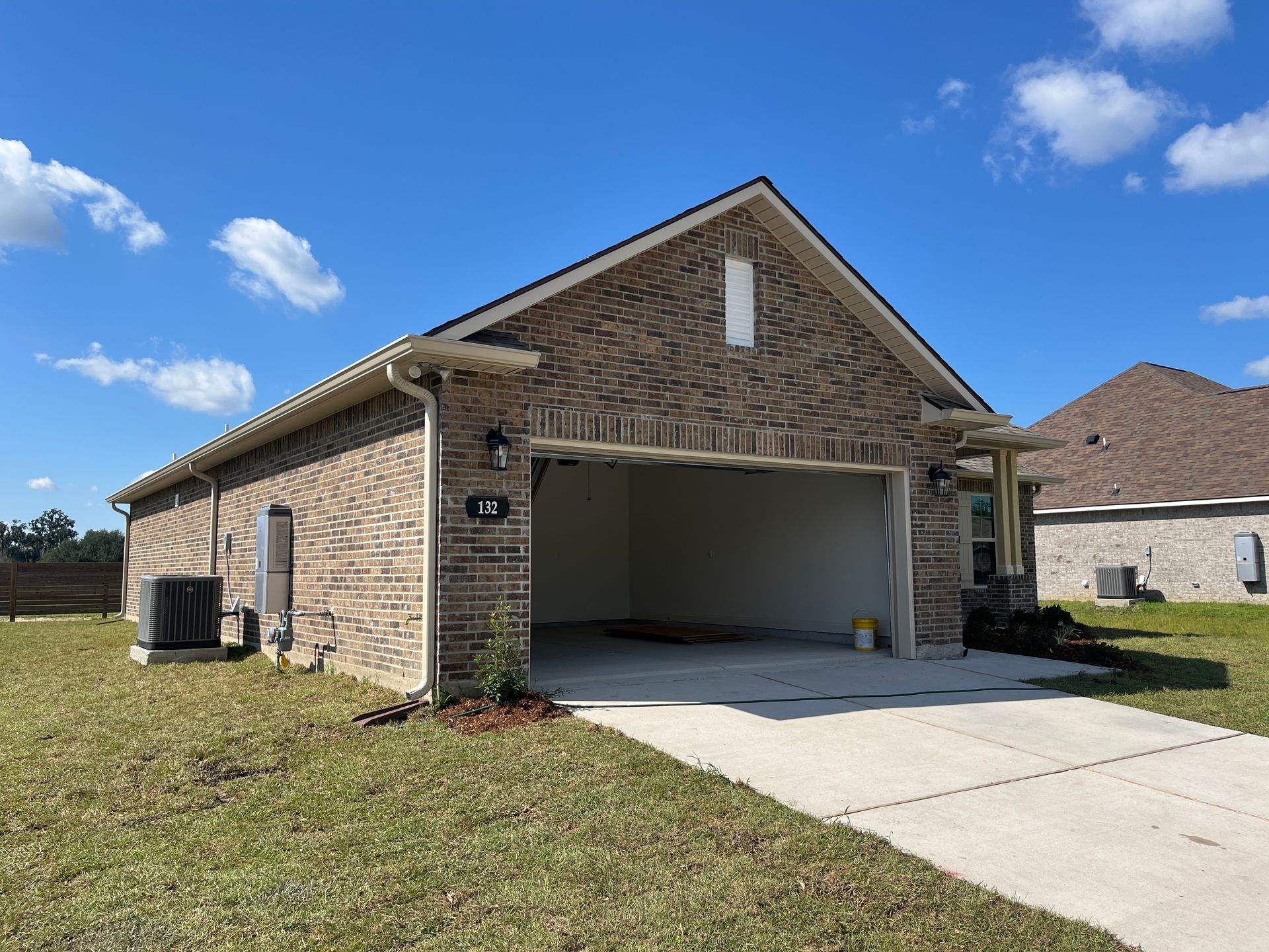 A single-story, tan brick house with an empty two-car garage, concrete driveway, and grassy yard under a blue sky.