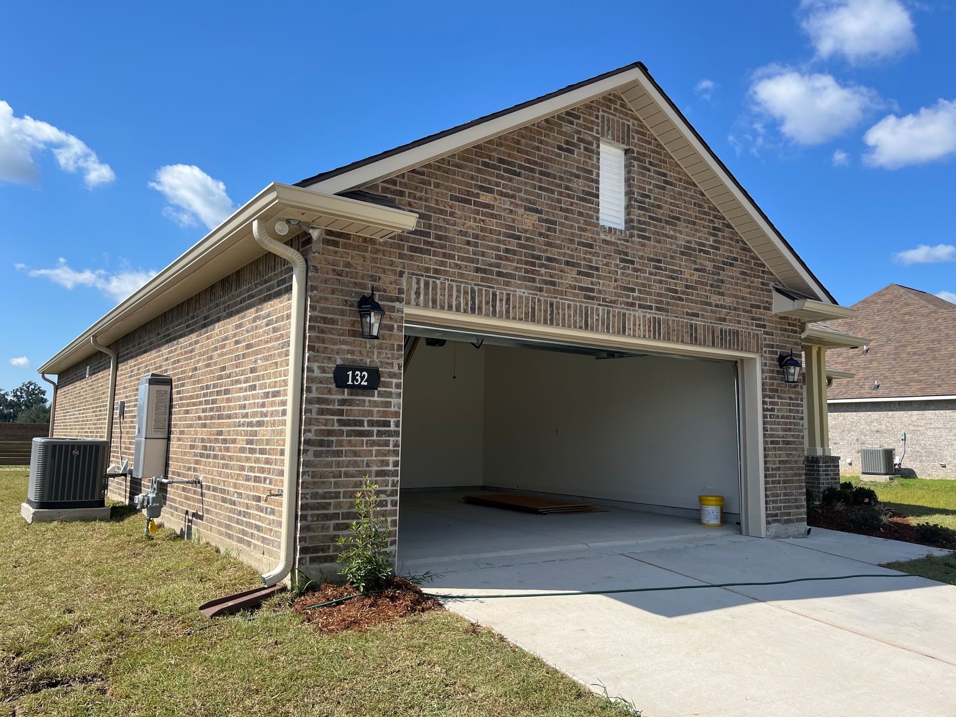 A tan brick detached garage with an open bay door, a white trim roof, and an air conditioning unit on a grassy lawn.
