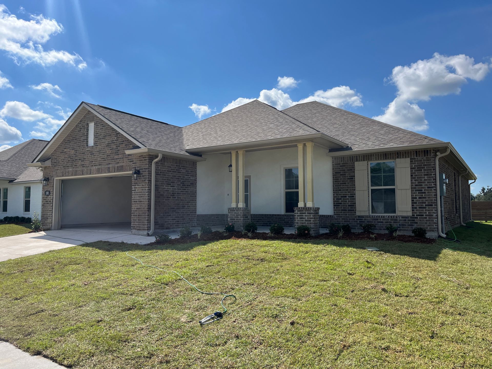 A single-story, tan stucco home with brick siding, a gray shingled roof, a two-car garage, and a front porch.