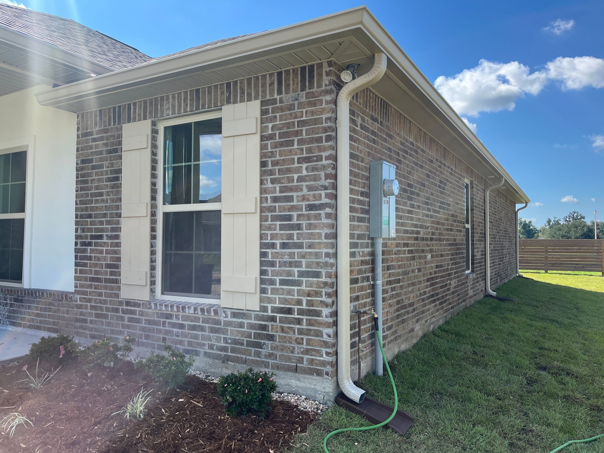 A side view of a residential house featuring brown brick walls, cream-colored window shutters, and a white gutter pipe.