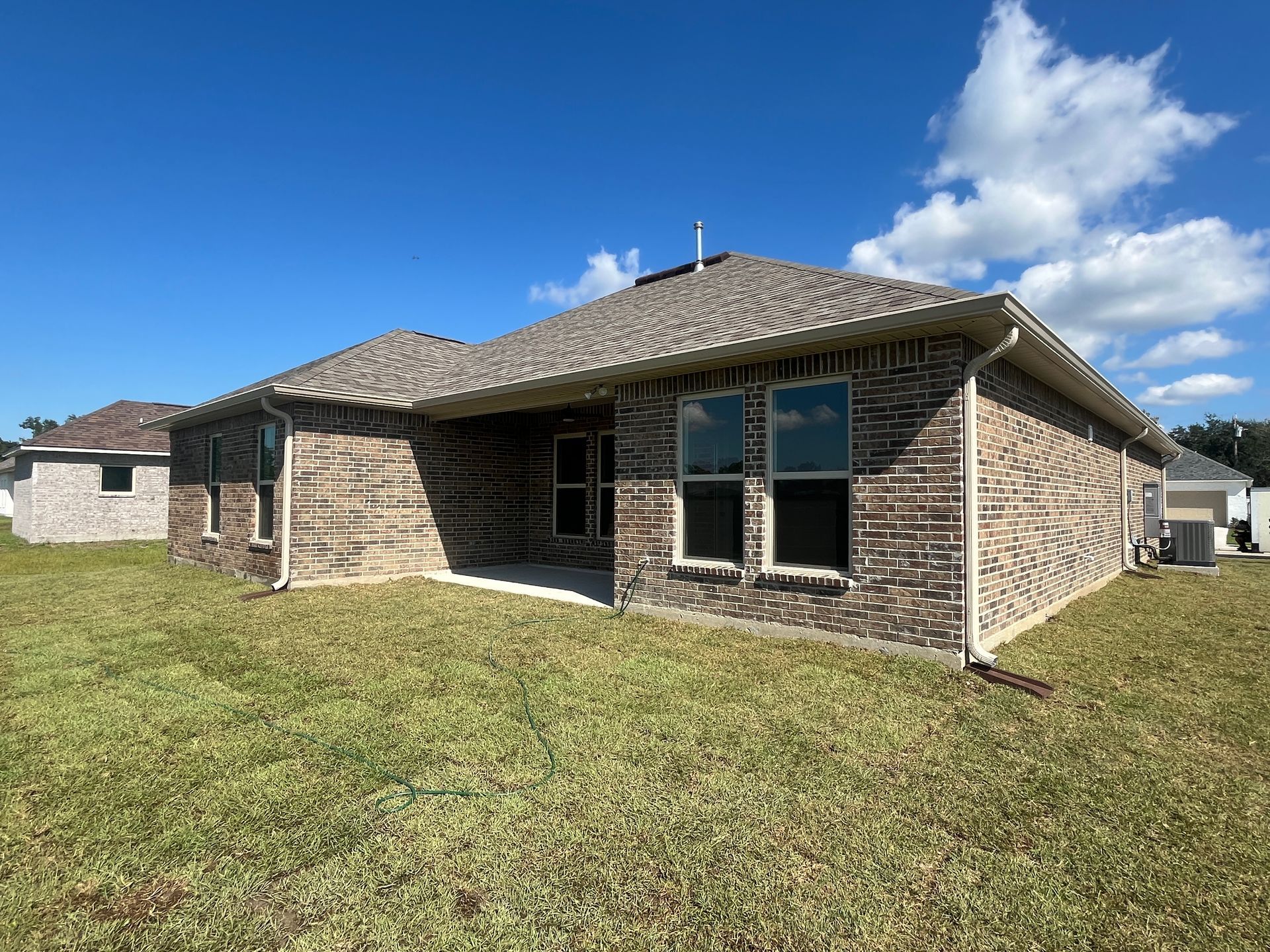 The back of a tan brick house with a covered patio, shingled roof, and a grassy lawn under a clear blue sky.