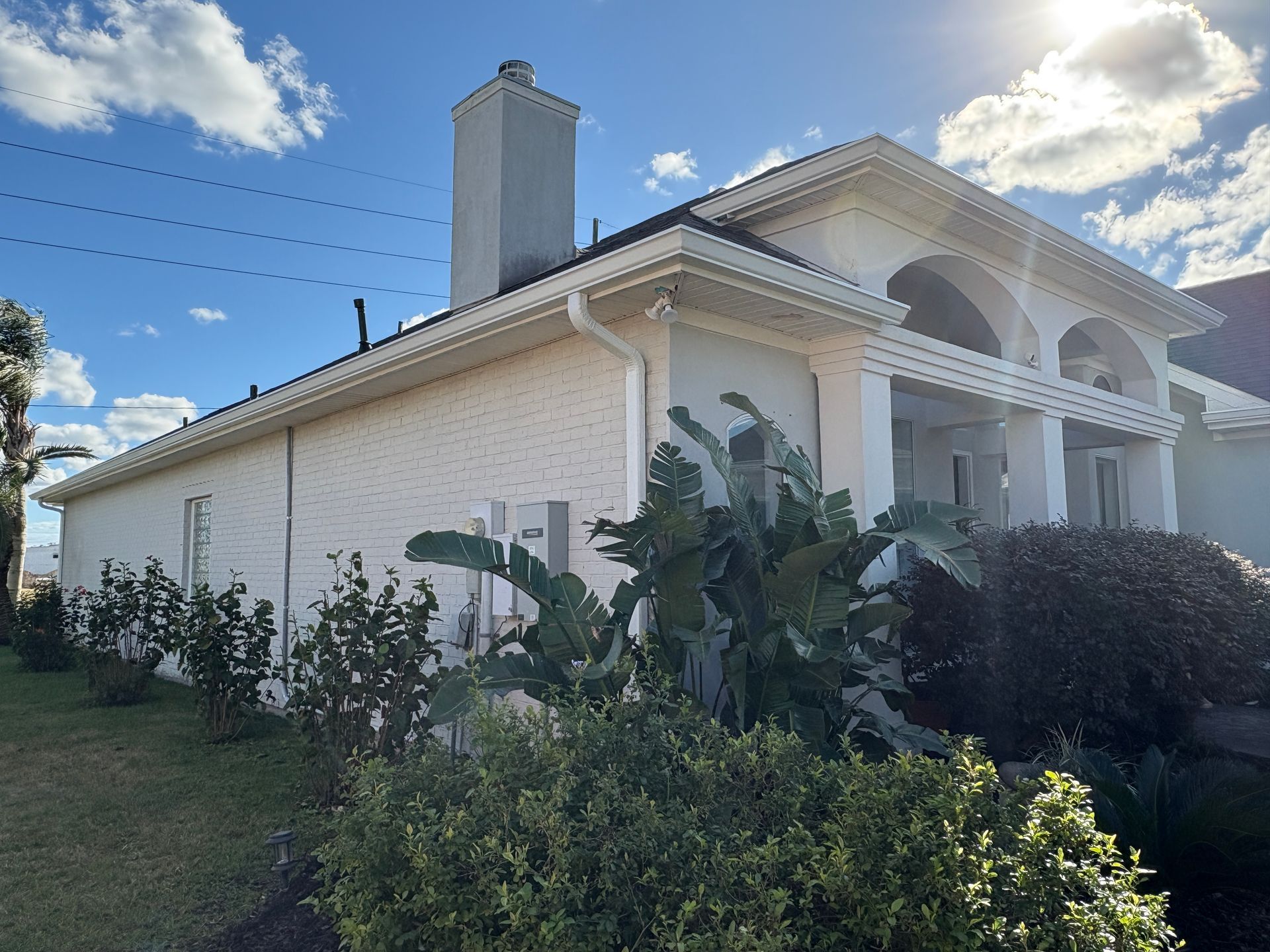A single-story white stucco home with a tall chimney, arched entryway, and lush green landscaping on a sunny day.