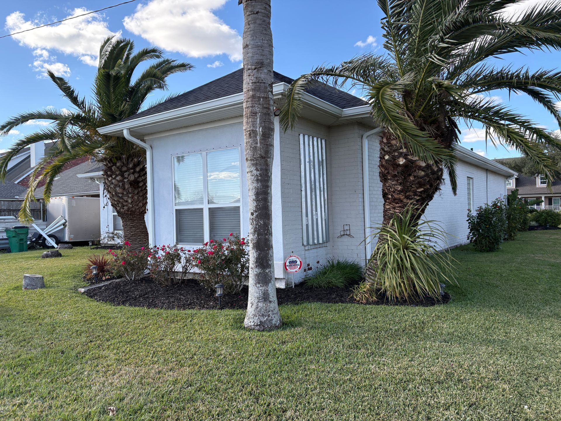 A white, single-story house with a grey roof, flanked by two large palm trees and surrounded by a grassy lawn.