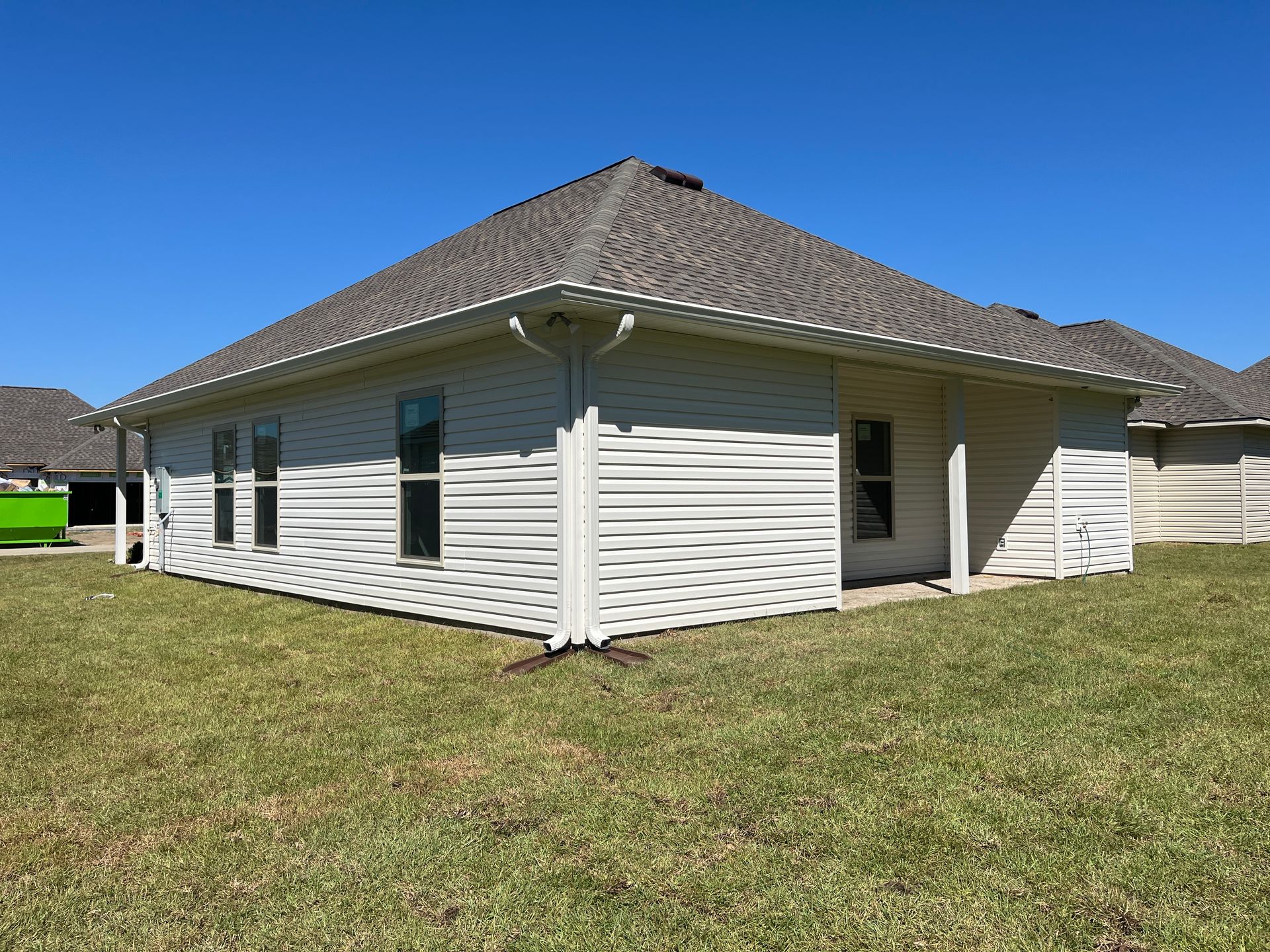 A side and rear view of a one-story, light-colored vinyl-sided house with a dark shingle roof, set on a sunny lawn.