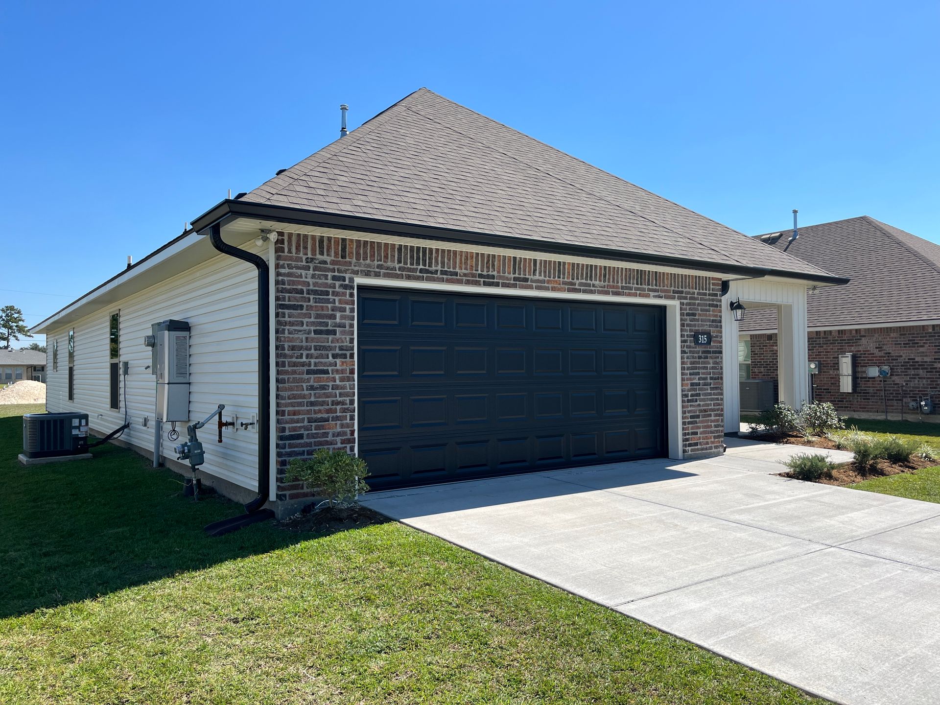 A single-story suburban house with a dark garage door, beige siding, brick facade, and a concrete driveway.