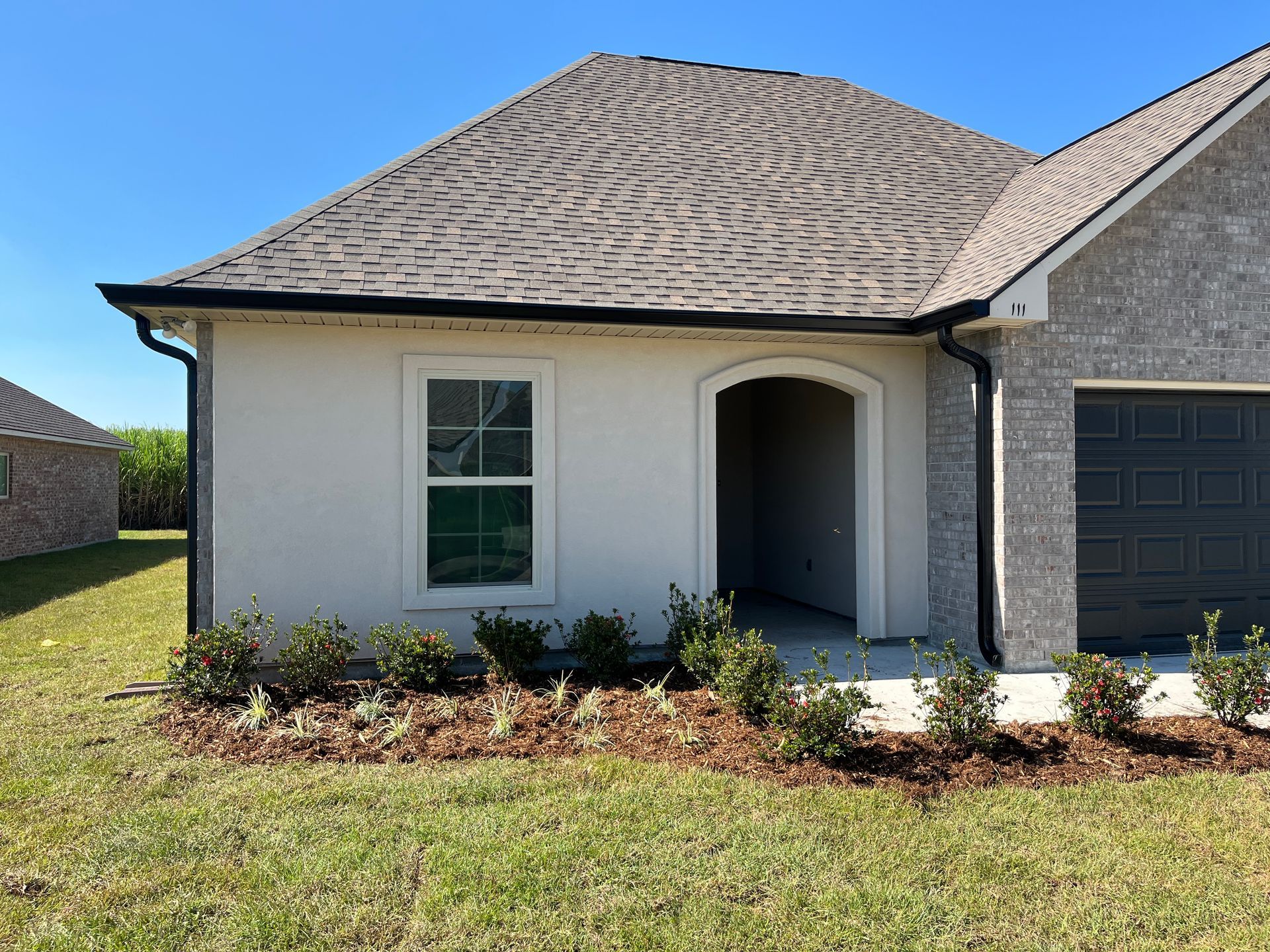 A single-story house exterior with light-colored stucco, a brick garage section, a gray shingle roof, and landscaping.