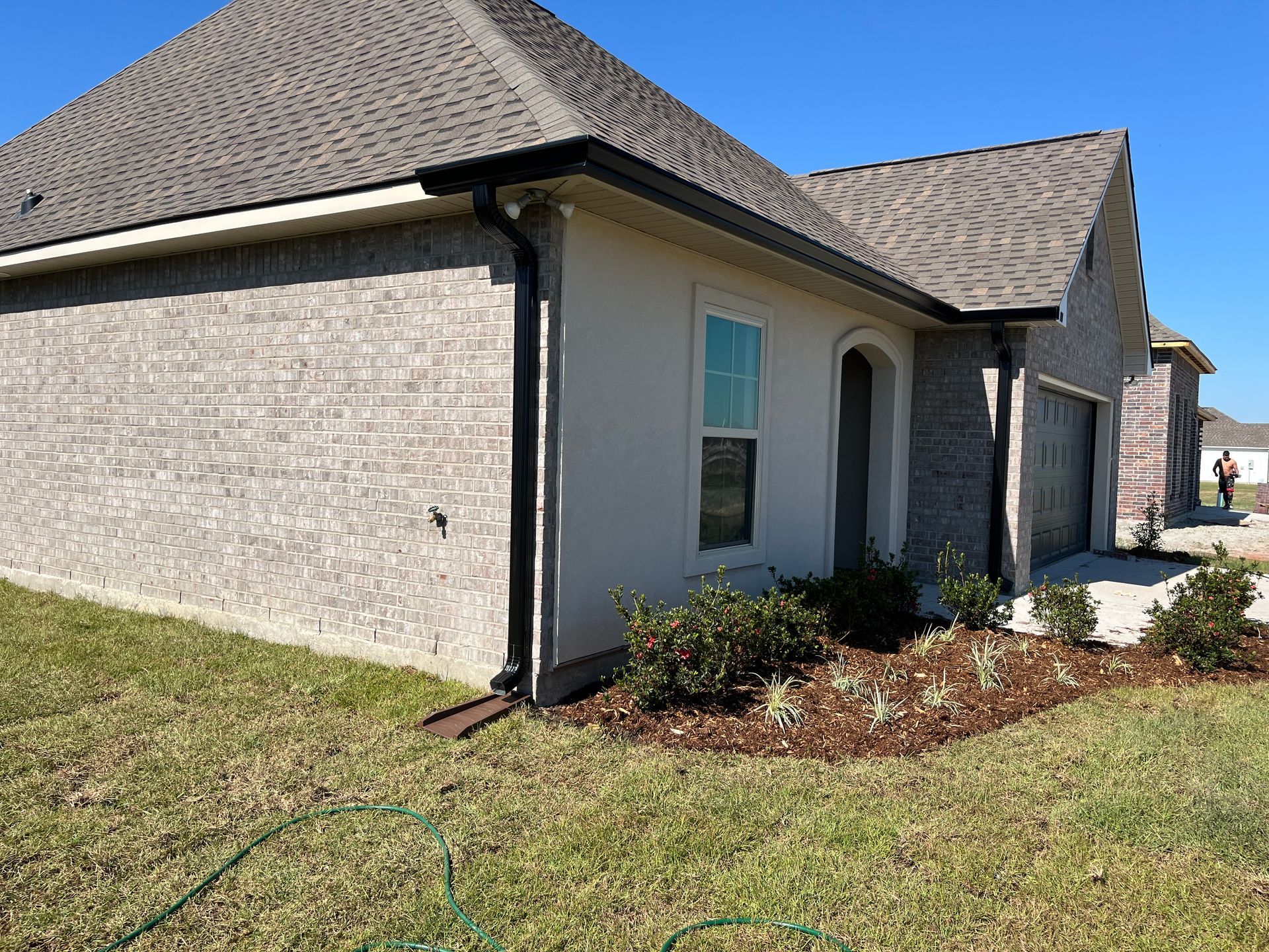 A tan brick house with a dark shingled roof, black gutters, and a garage under a clear blue sky.