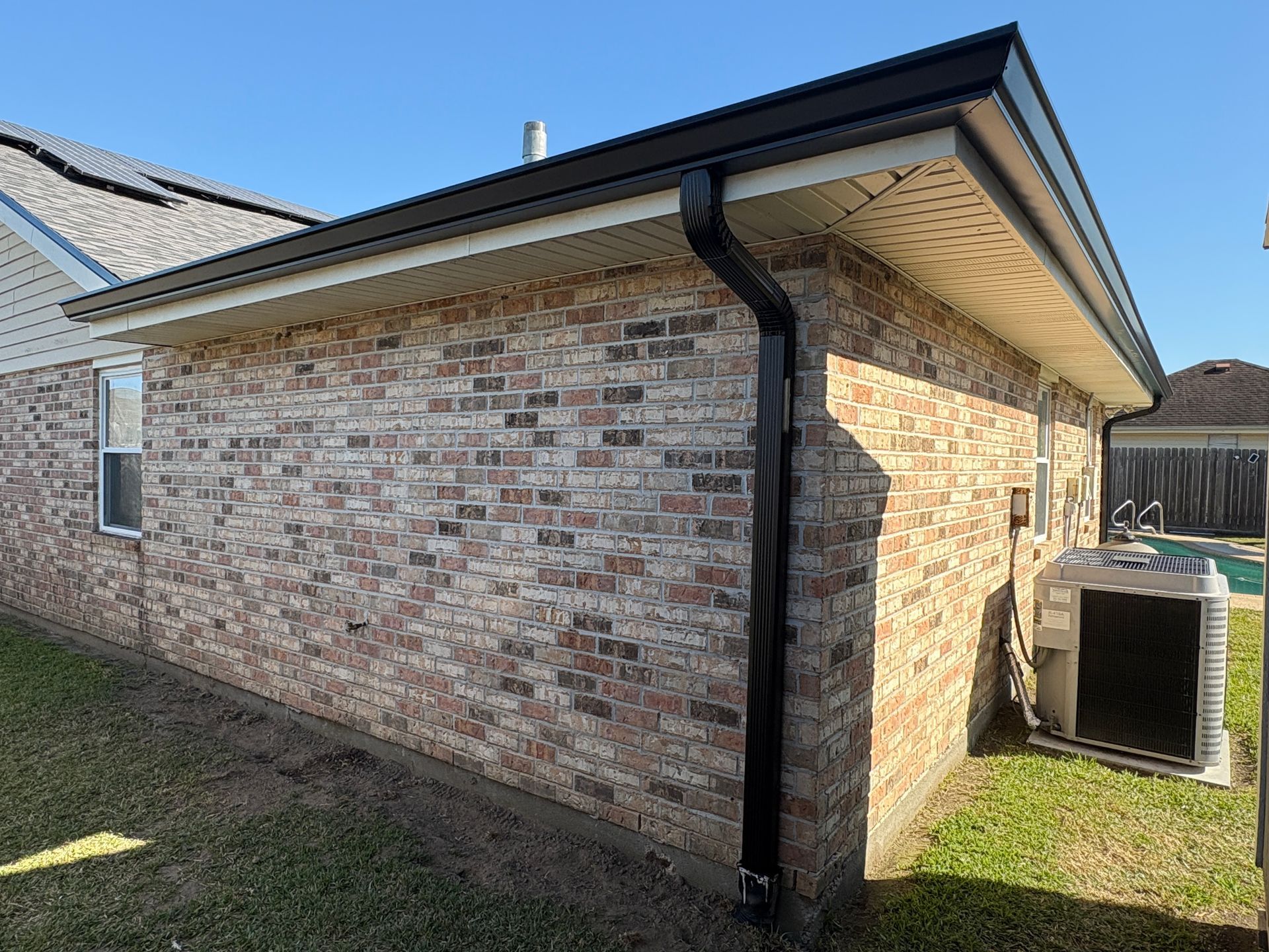A side view of a brick house with a black downspout, an HVAC unit, and a small patch of grass under a clear blue sky.