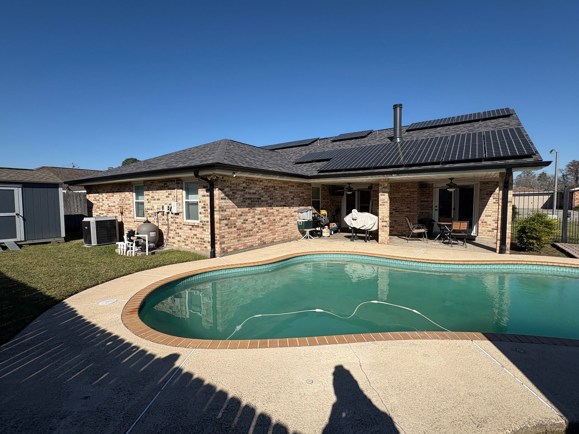 A brick house with a dark shingled roof overlooks a backyard swimming pool under a clear blue sky.