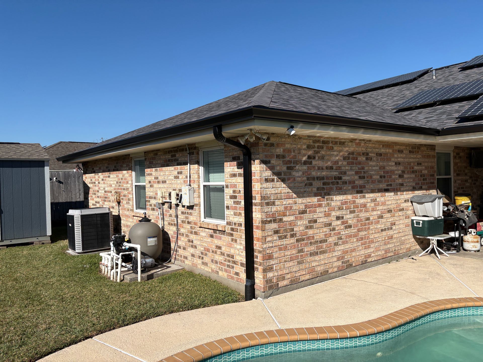 The brick exterior wall of a house with a shingled roof, pool equipment, and an adjacent backyard pool under a blue sky.