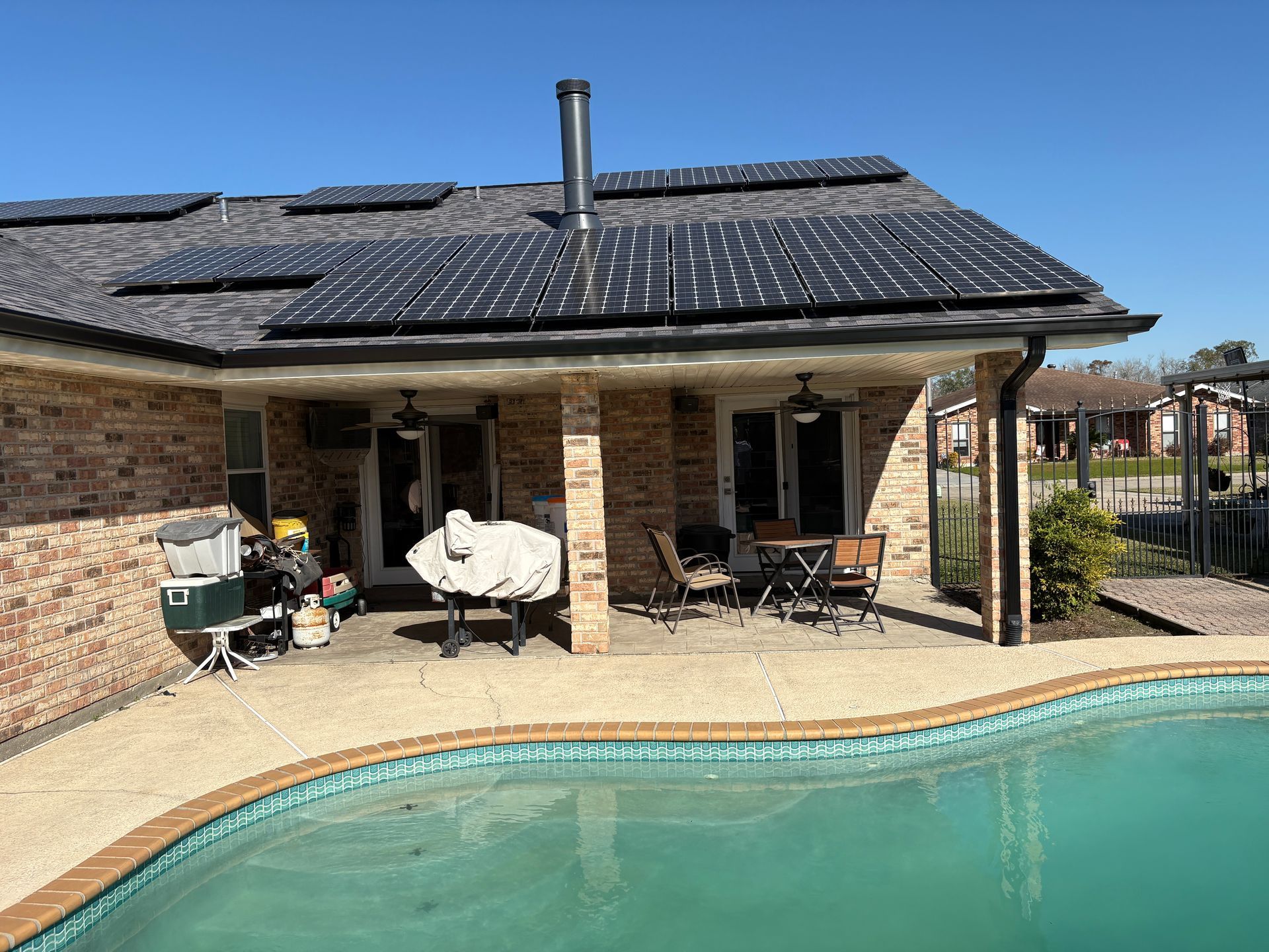 Solar panels cover the roof of a brick house with a covered patio, outdoor furniture, and a swimming pool in the yard.