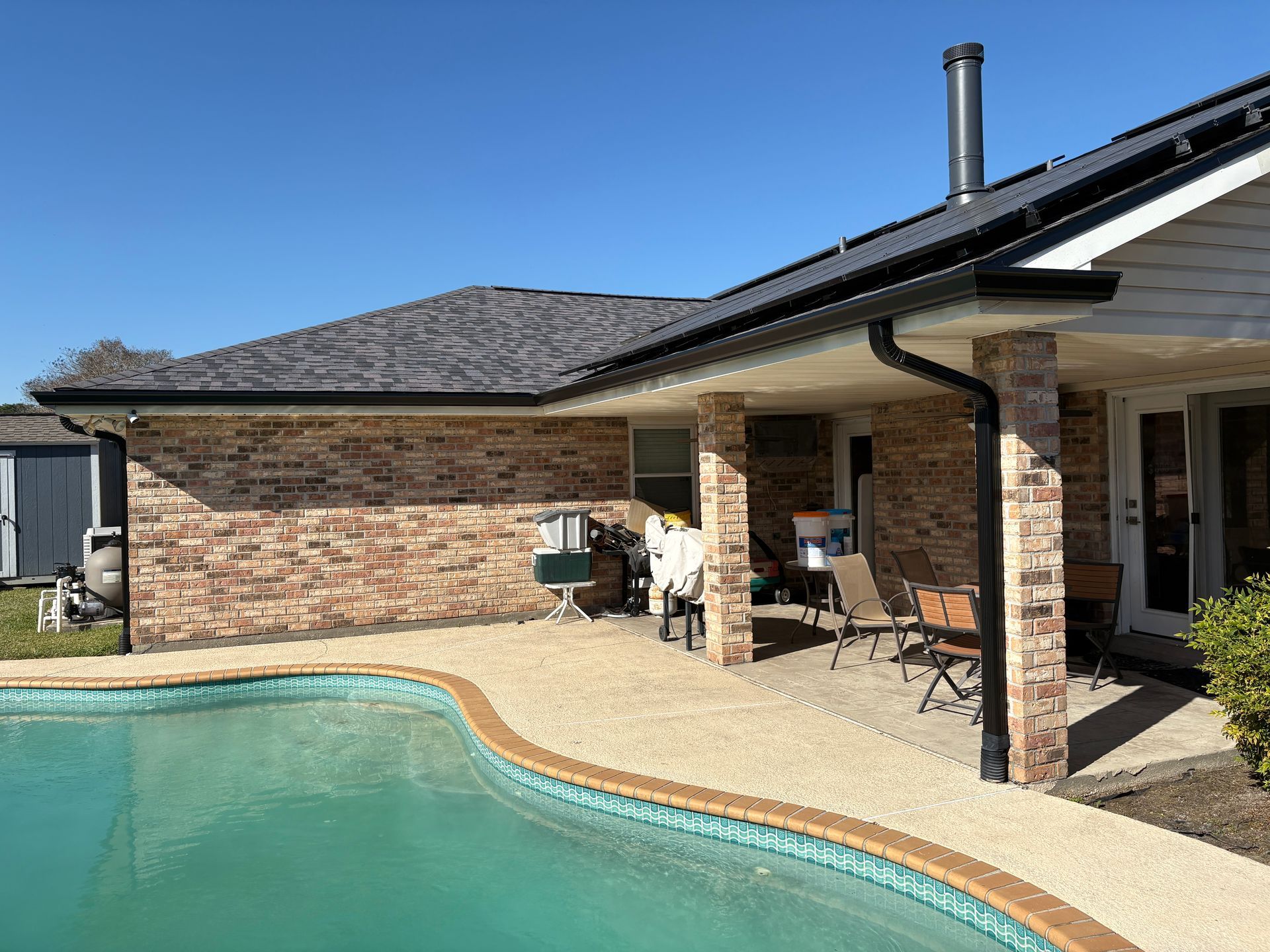 A swimming pool beside a brick house with a covered patio, outdoor furniture, and a clear blue sky.