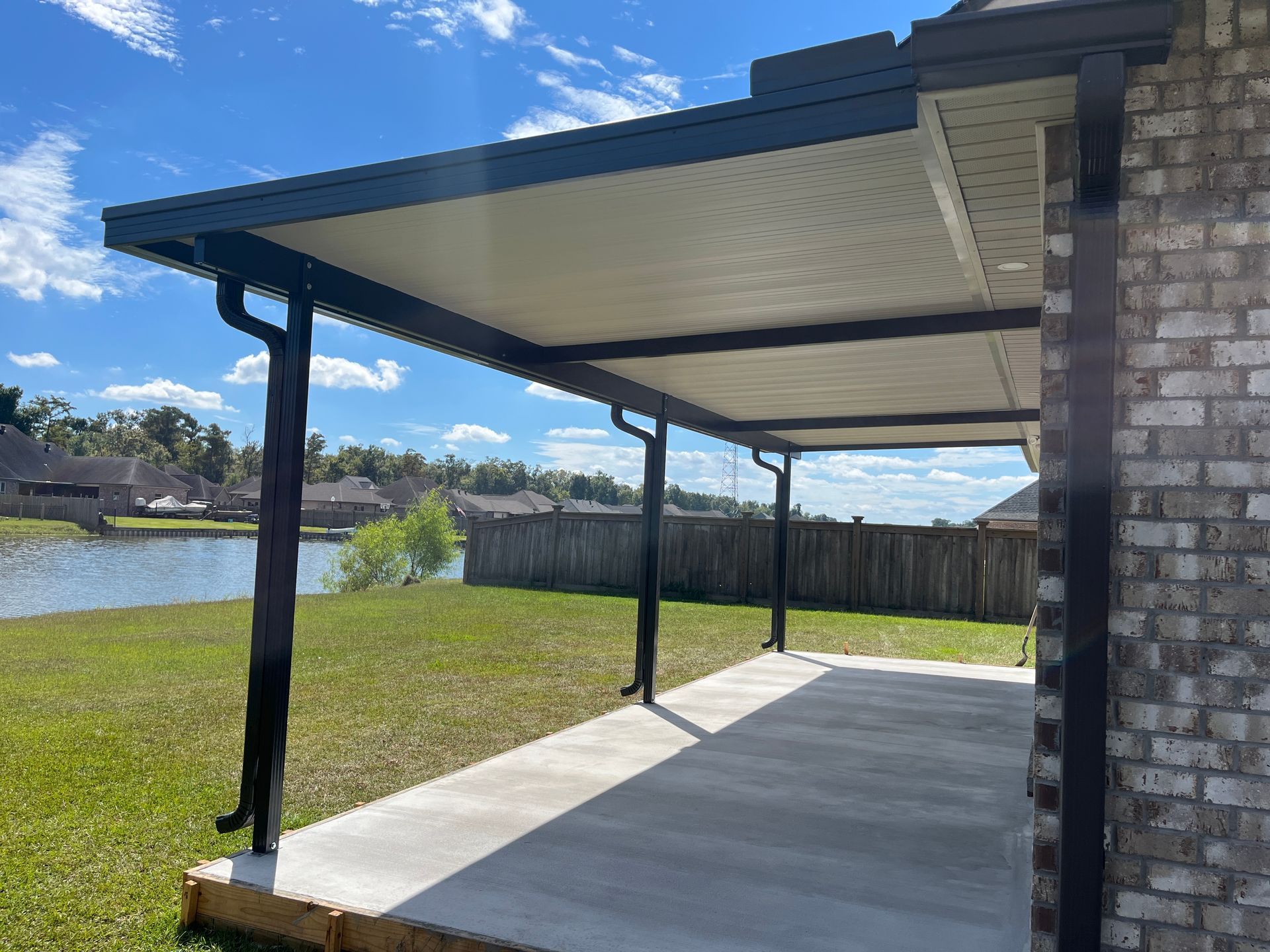 A covered patio with a dark metal frame and off-white ceiling extending from a brick house, overlooking a grassy yard.