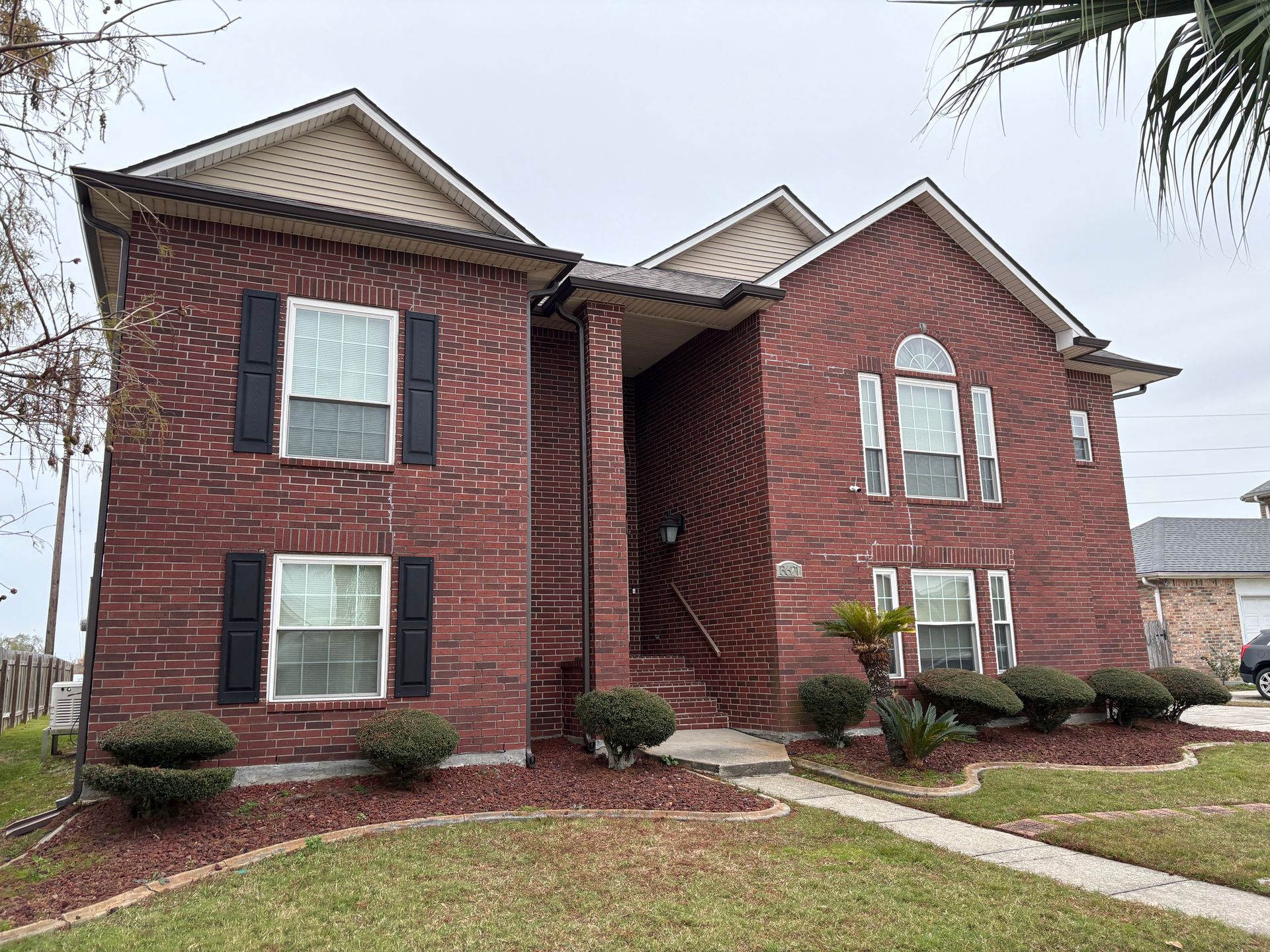 A two-story red brick suburban house with a recessed entry, dark shutters, and a landscaped front yard under a gray sky.