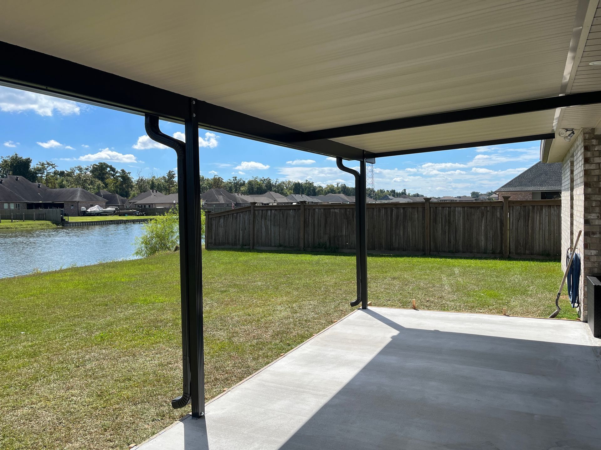 A covered concrete patio overlooking a backyard with a pond, grass, and a wooden fence under a blue sky.