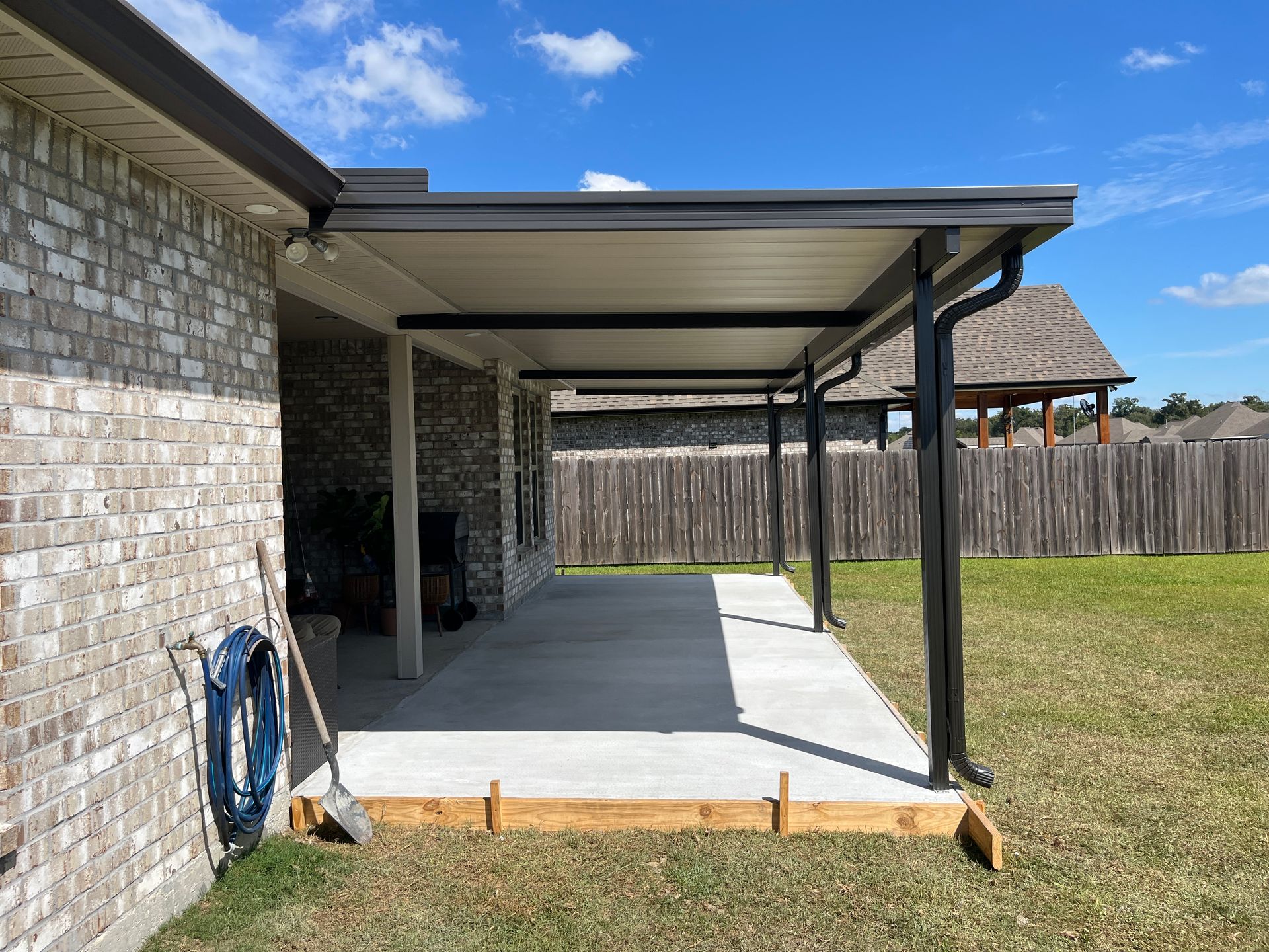 A brick house with a newly poured concrete patio extension under a dark metal roof overhang in a sunny backyard.