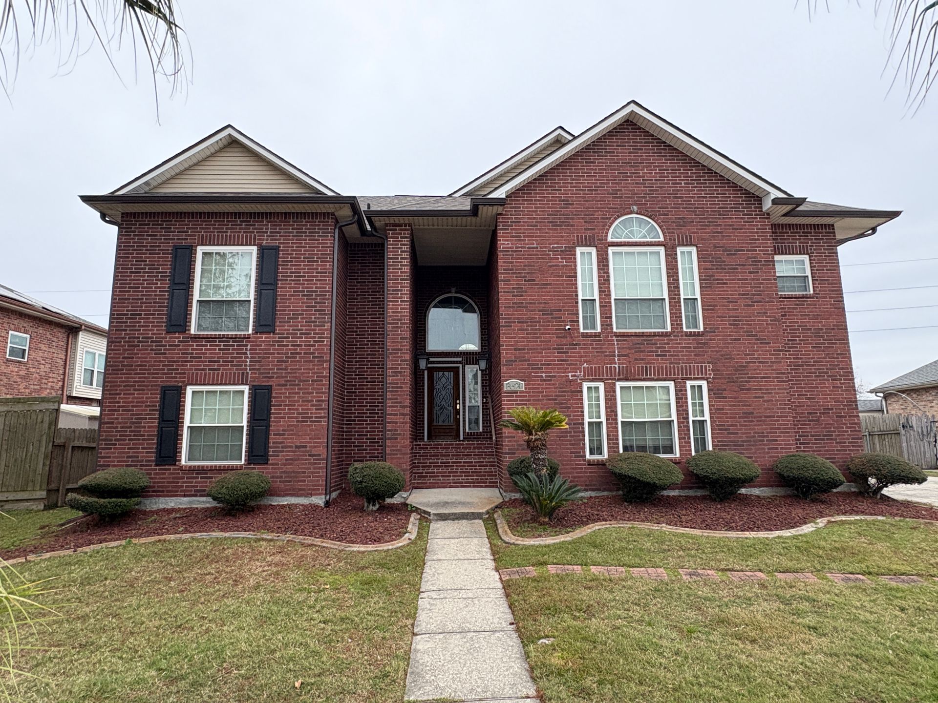 Two-story red brick house with a dark roof, black shutters, white window trim, and a centered front door with arched glass.
