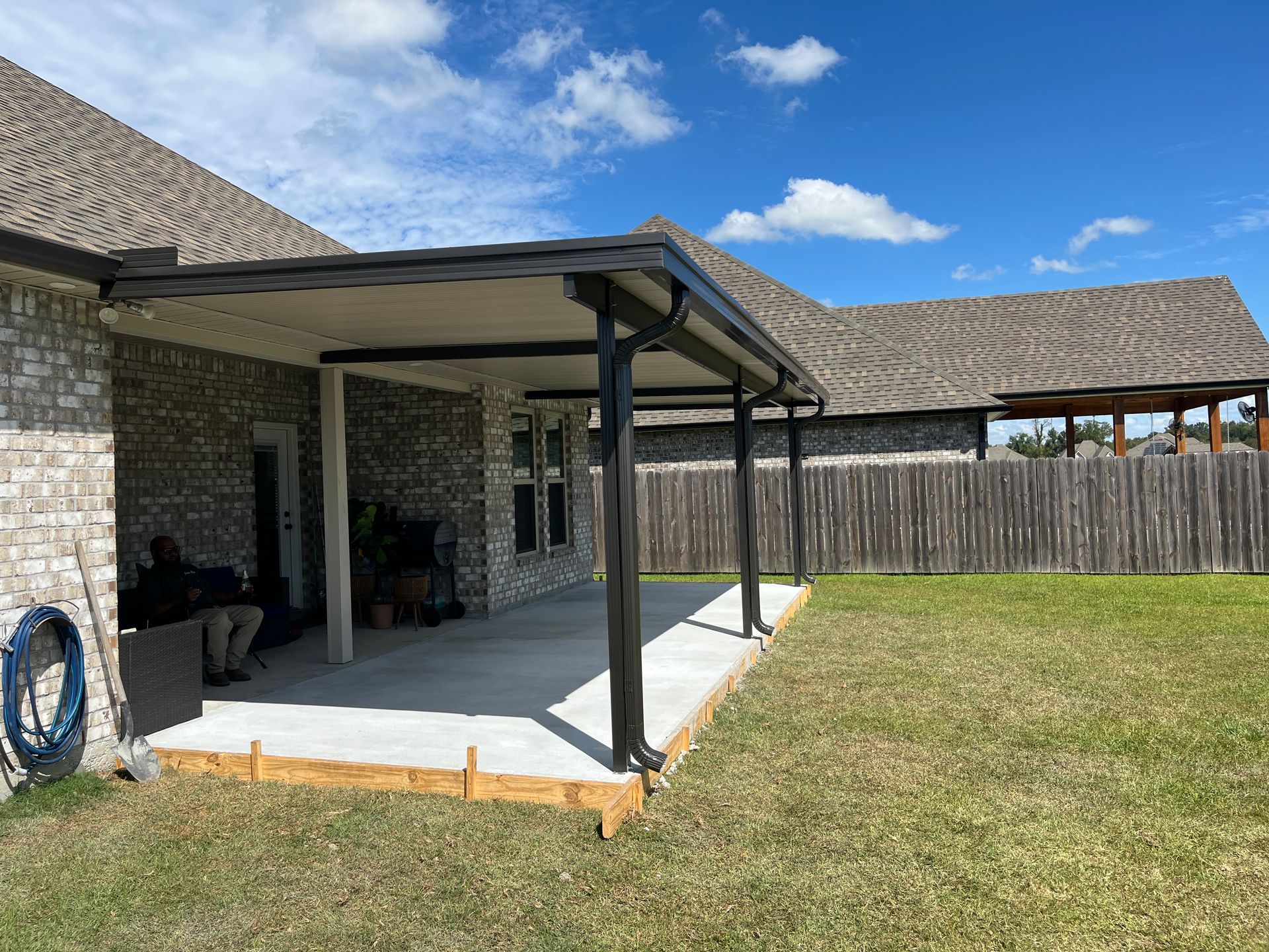 A covered stone patio with a concrete floor, black support beams, and a dark roof extension in a backyard.