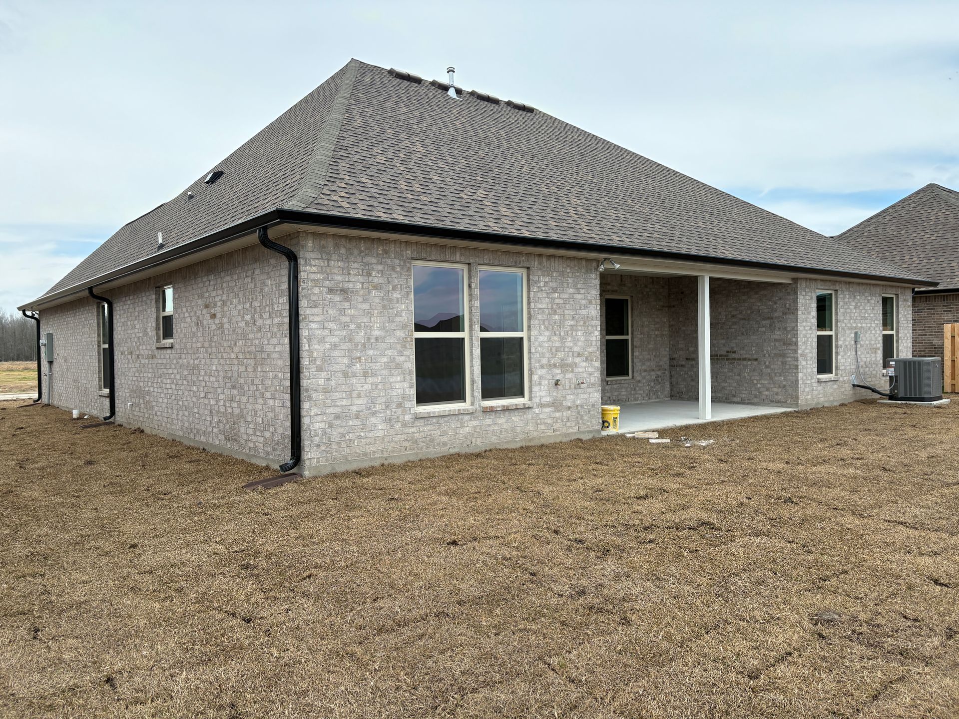 The back view of a light brick house featuring a covered patio, a gray shingled hip roof, and an unfinished yard.