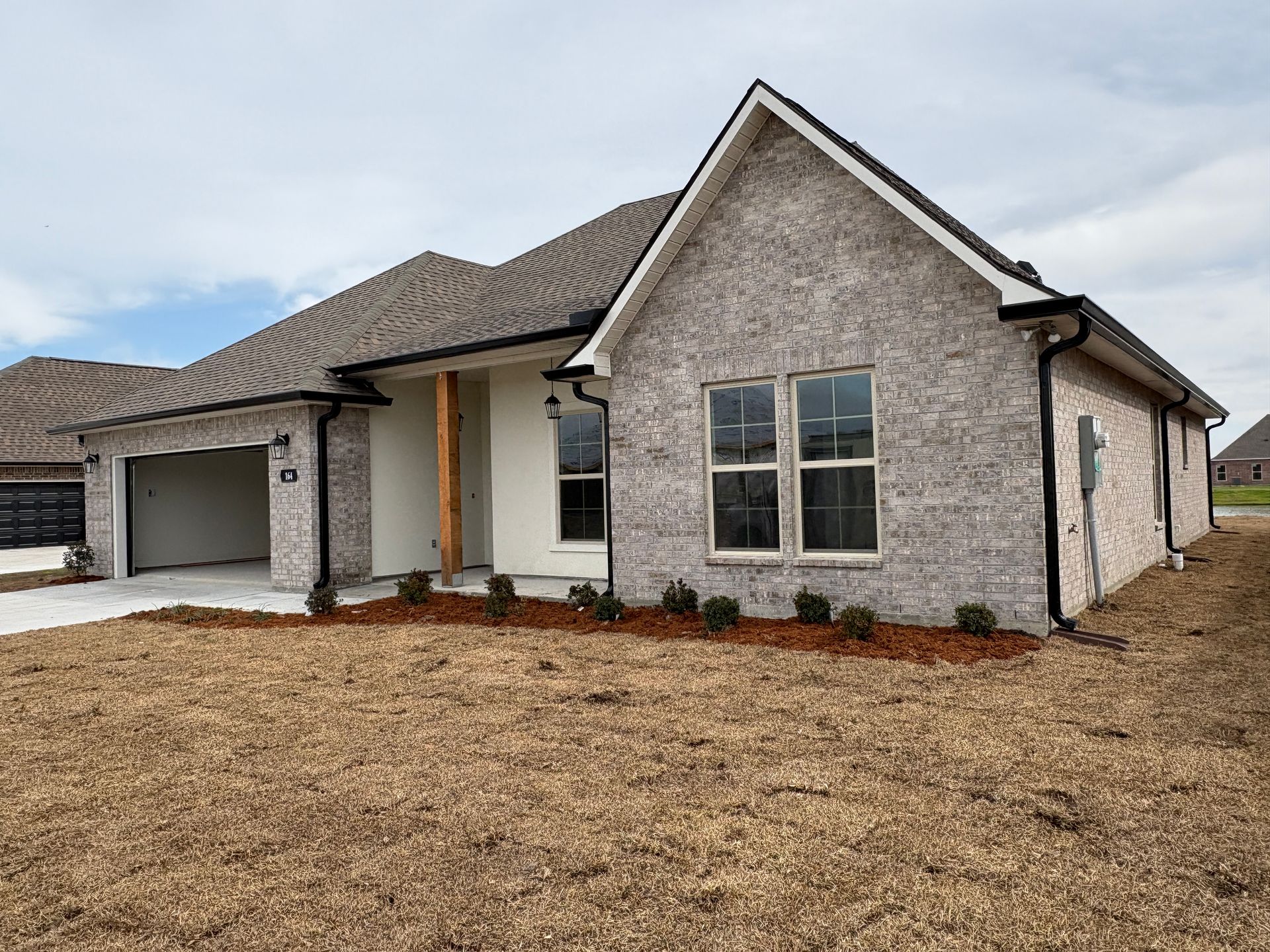 A single-story, light-grey brick house with a dark shingle roof, a two-car garage, and a wood-post covered front porch.